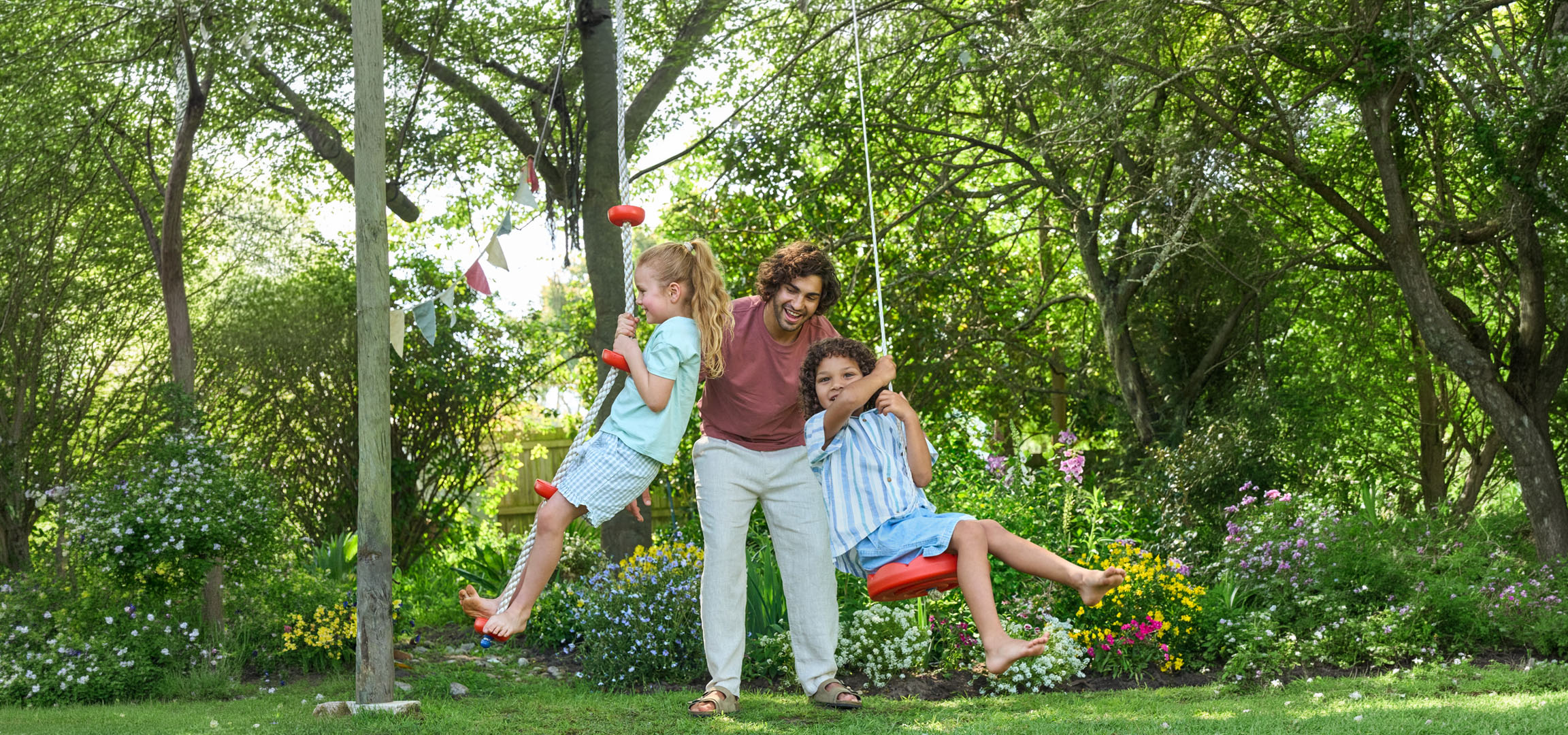 Father and two children playing on swings in a lush green garden.