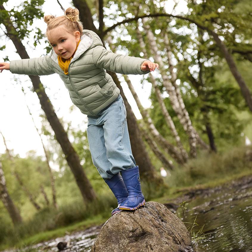 Little girl in a puffer jacket, jeans, and wellington boots balancing on a rock in the woods.