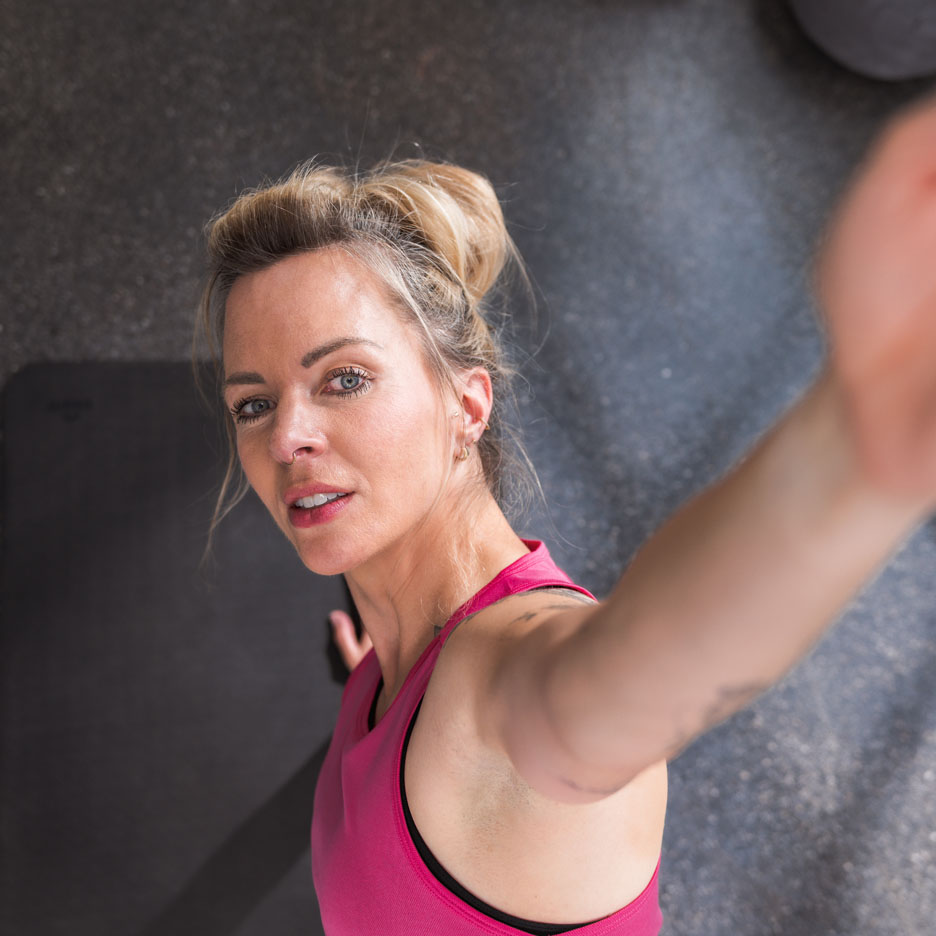 A woman in a pink athletic top takes a selfie at the gym.