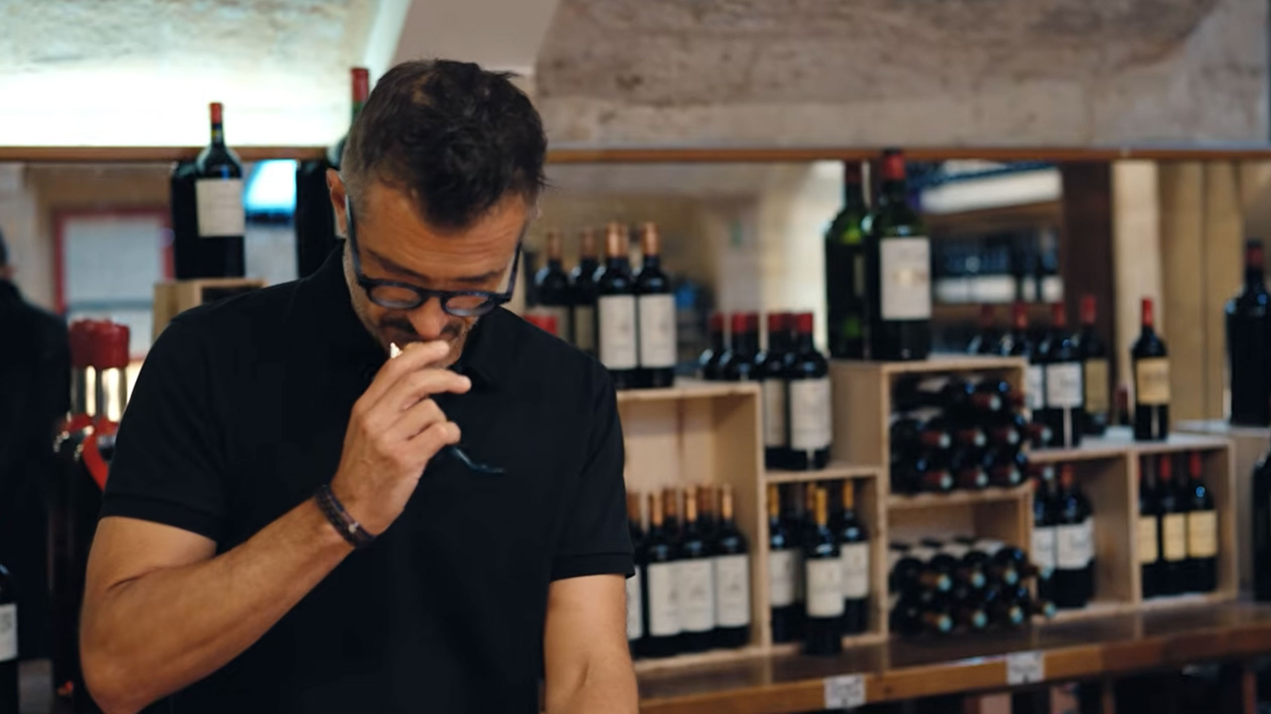 Man in a wine cellar tasting wine, surrounded by bottles on wooden shelves.