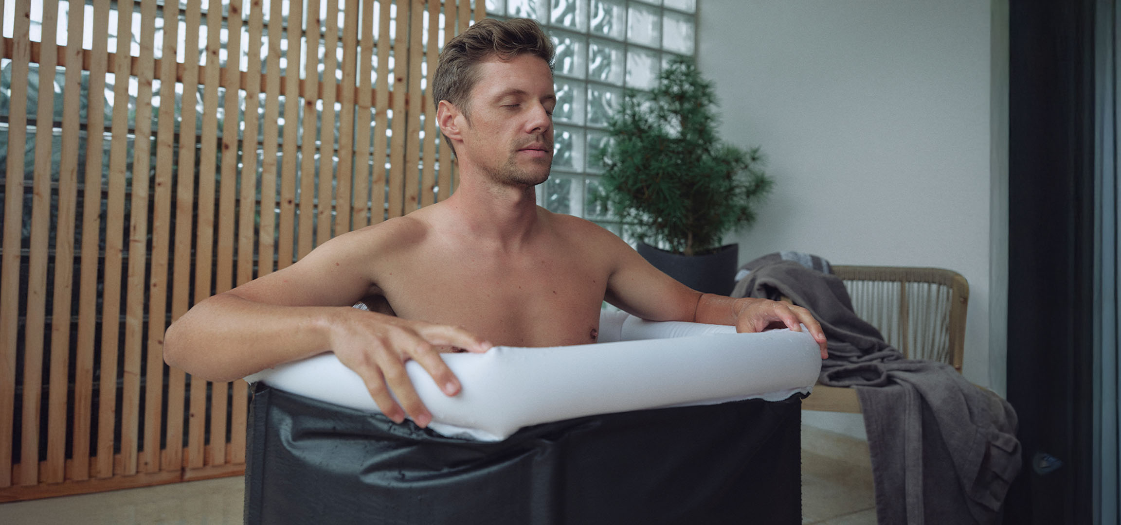 Man in a black ice bath tub on a balcony with wooden paneling and glass doors.