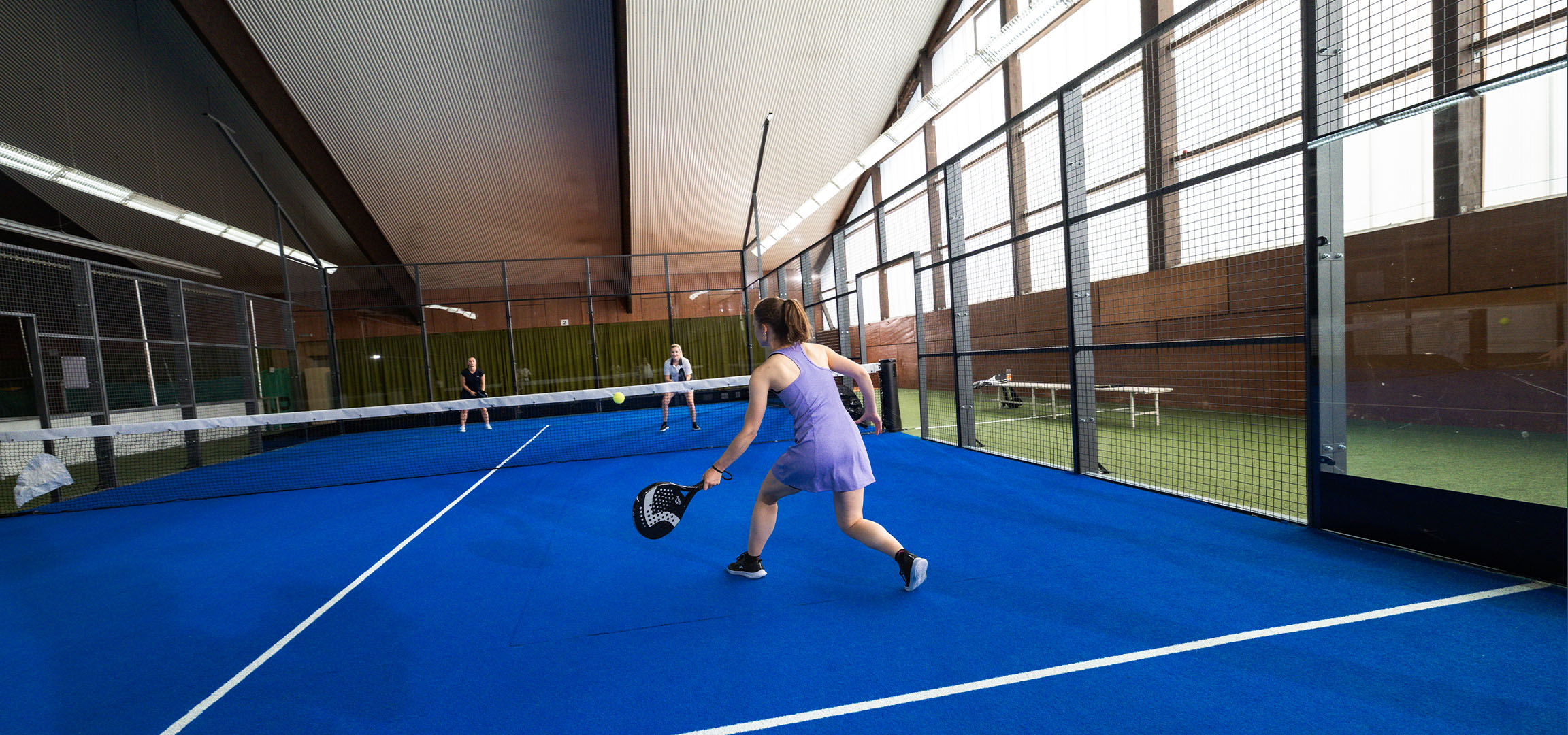 Woman playing padel on a blue court, with other players in the background.