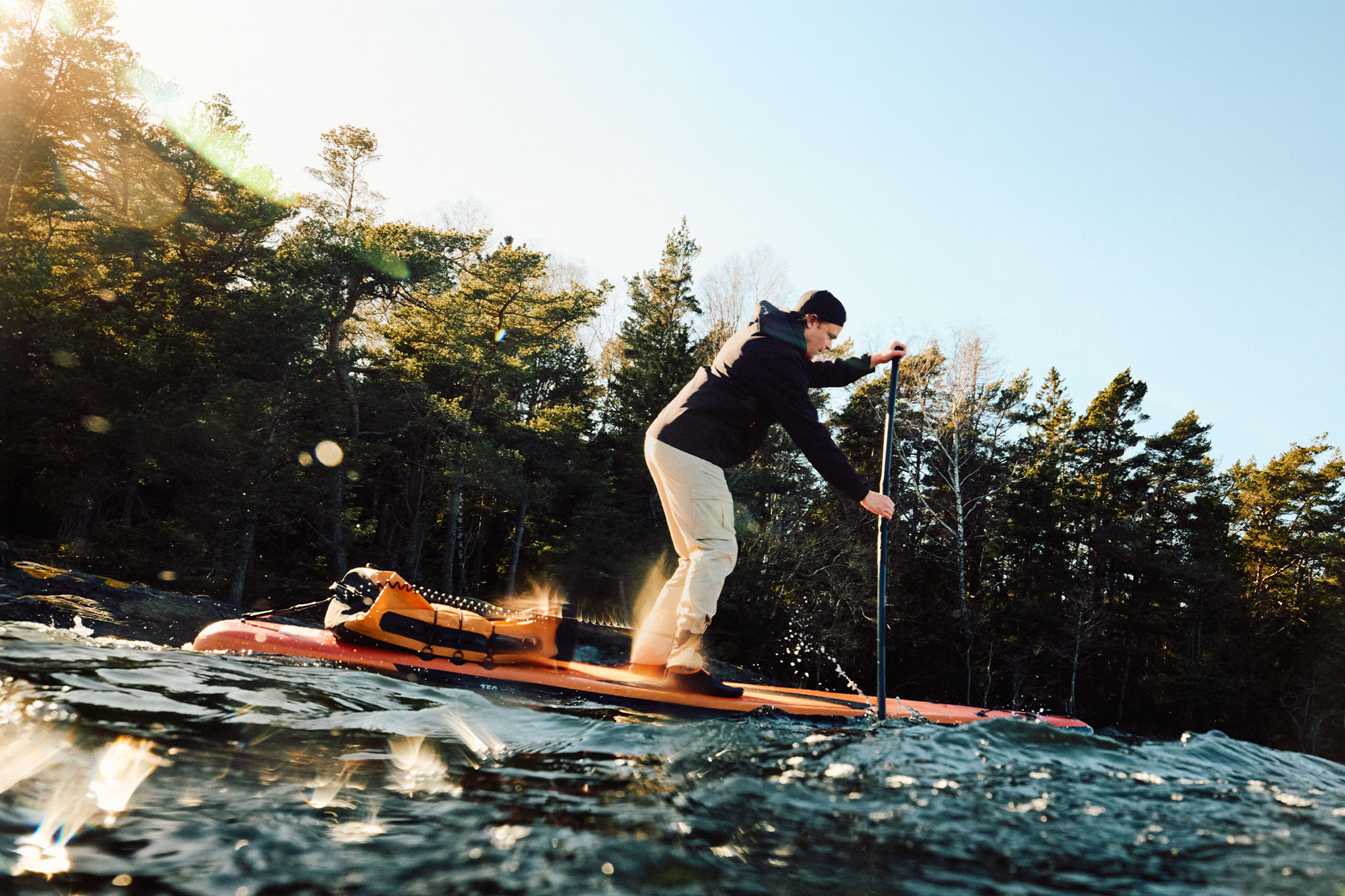 Άνδρας κάνει paddleboard με μια πορτοκαλί αδιάβροχη τσάντα δίπλα σε ένα δάσος.