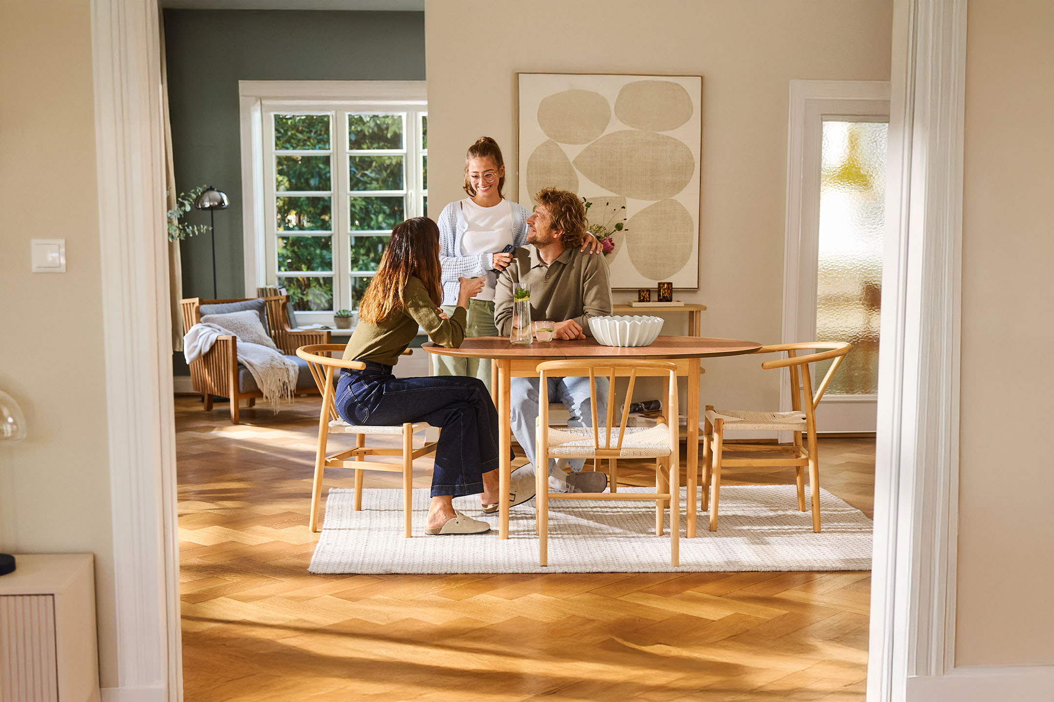 Three people chat at a wooden table in a bright living room with large windows.