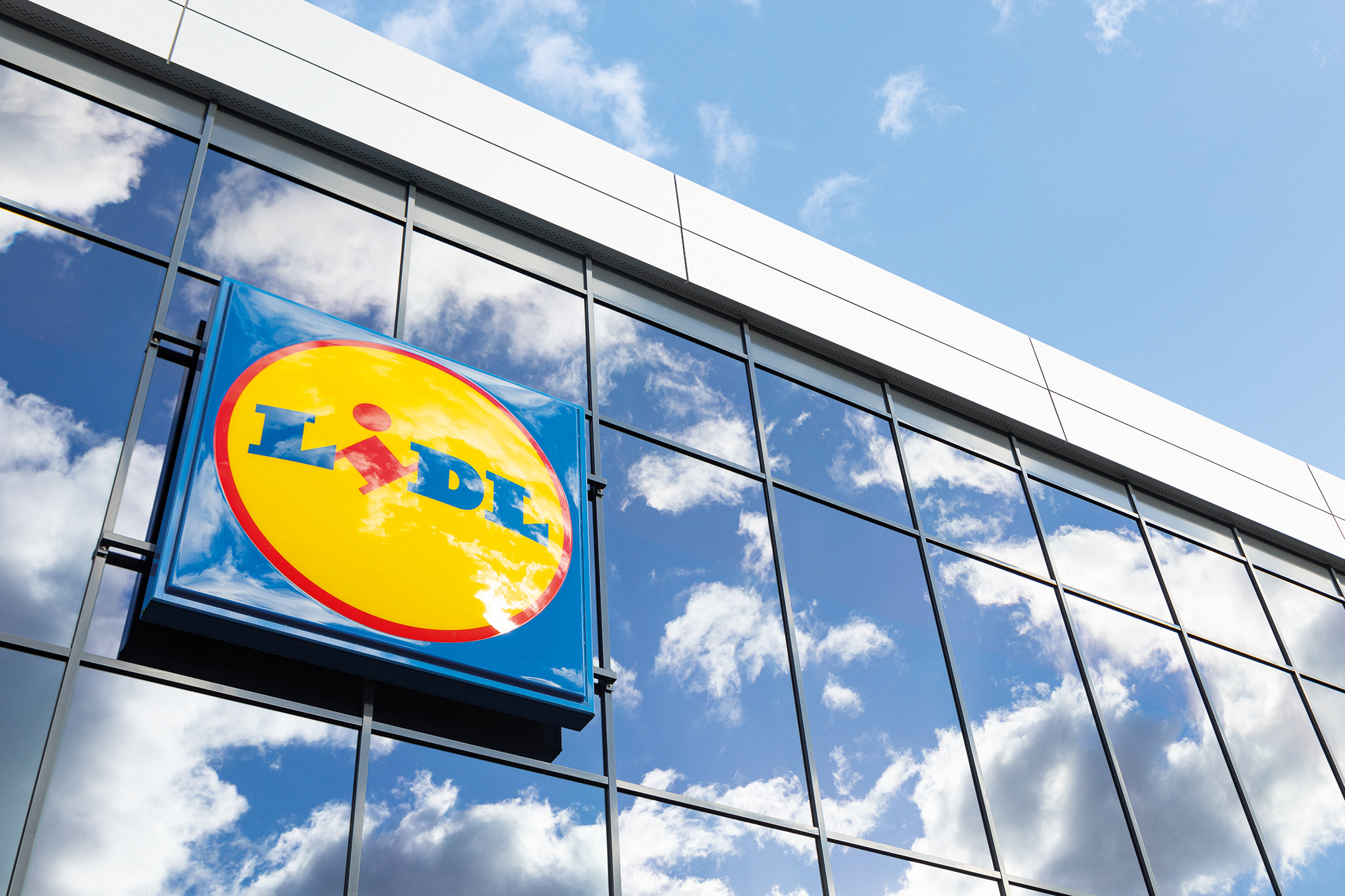 Lidl sign on a glass building facade, with sky and clouds reflected.