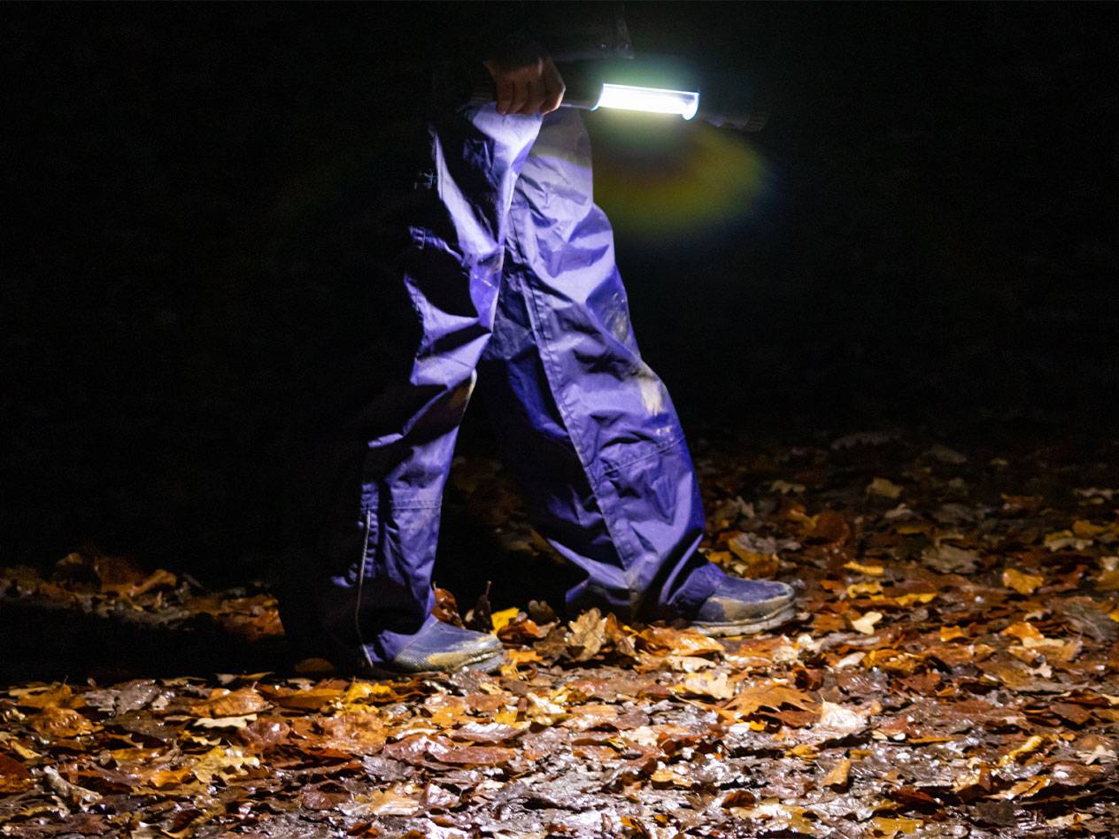 Person in blue waterproof trousers walking at night, illuminating fallen leaves with a flashlight.