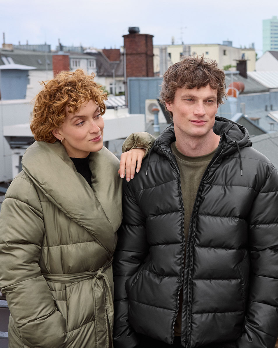 Two people in puffer jackets on a rooftop, woman in olive green, man in black.