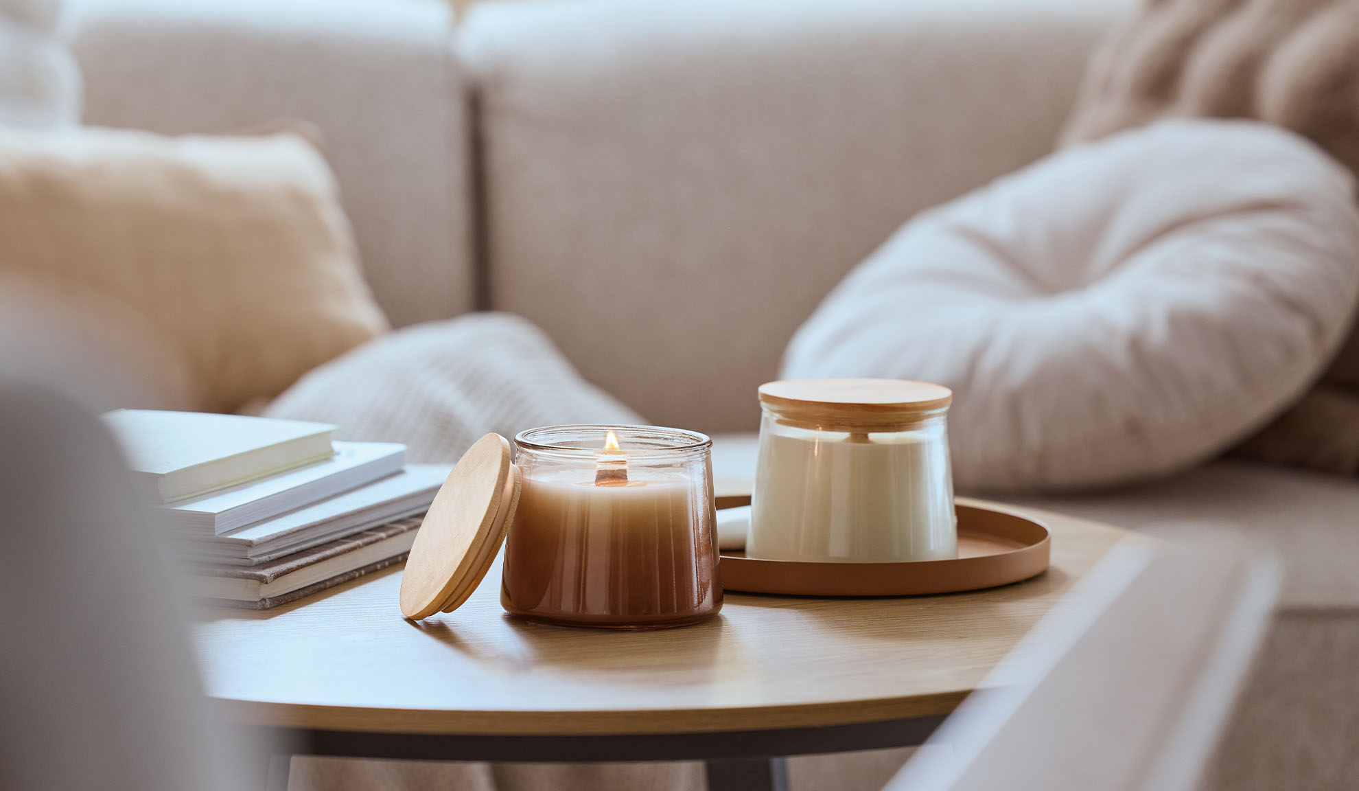 Δύο αρωματικά κεριά σε ξύλινο τραπέζι, το ένα αναμμένο, με έναν άνετο καναπέ στο παρασκήνιο.