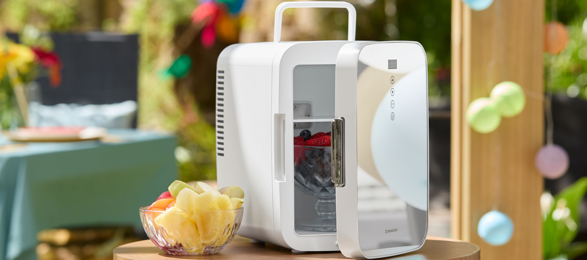 White mini cooler with open door filled with berries and a bowl of fruit on an outdoor table.