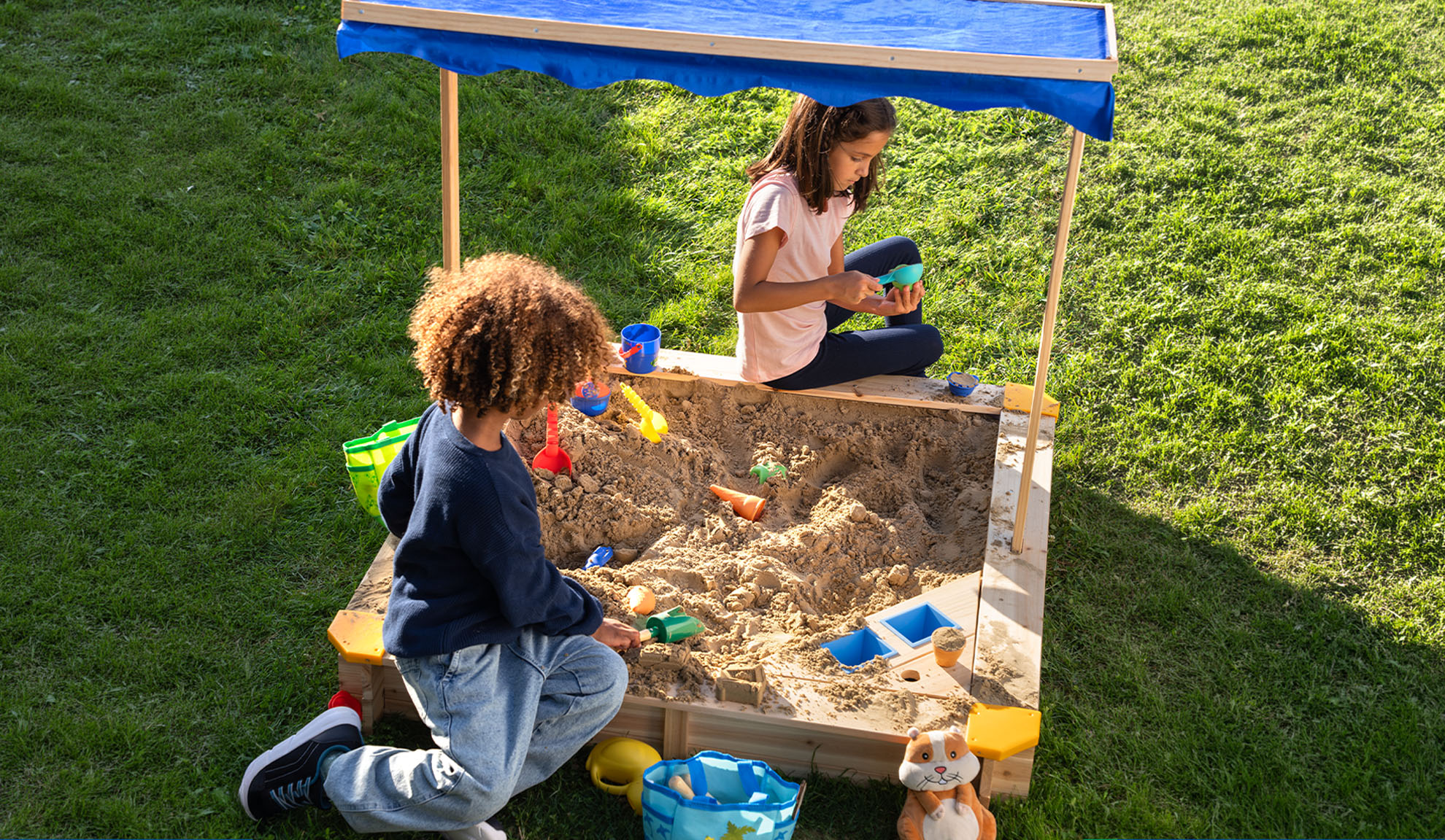 Two children playing in a sandbox with a blue canopy and sand toys.