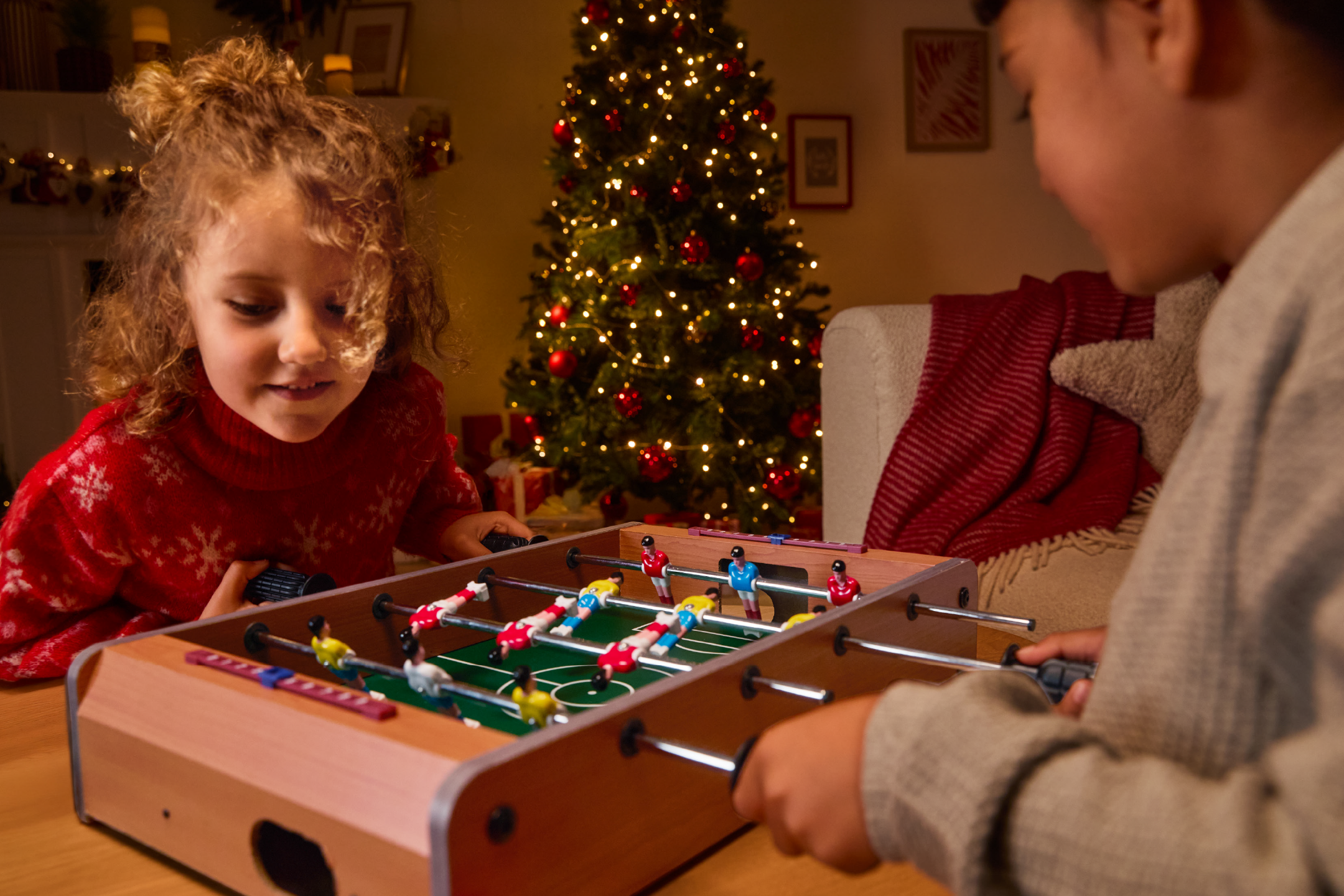 Δύο παιδιά παίζουν ποδοσφαιράκι σε ένα γιορτινό σαλόνι με χριστουγεννιάτικο δέντρο.