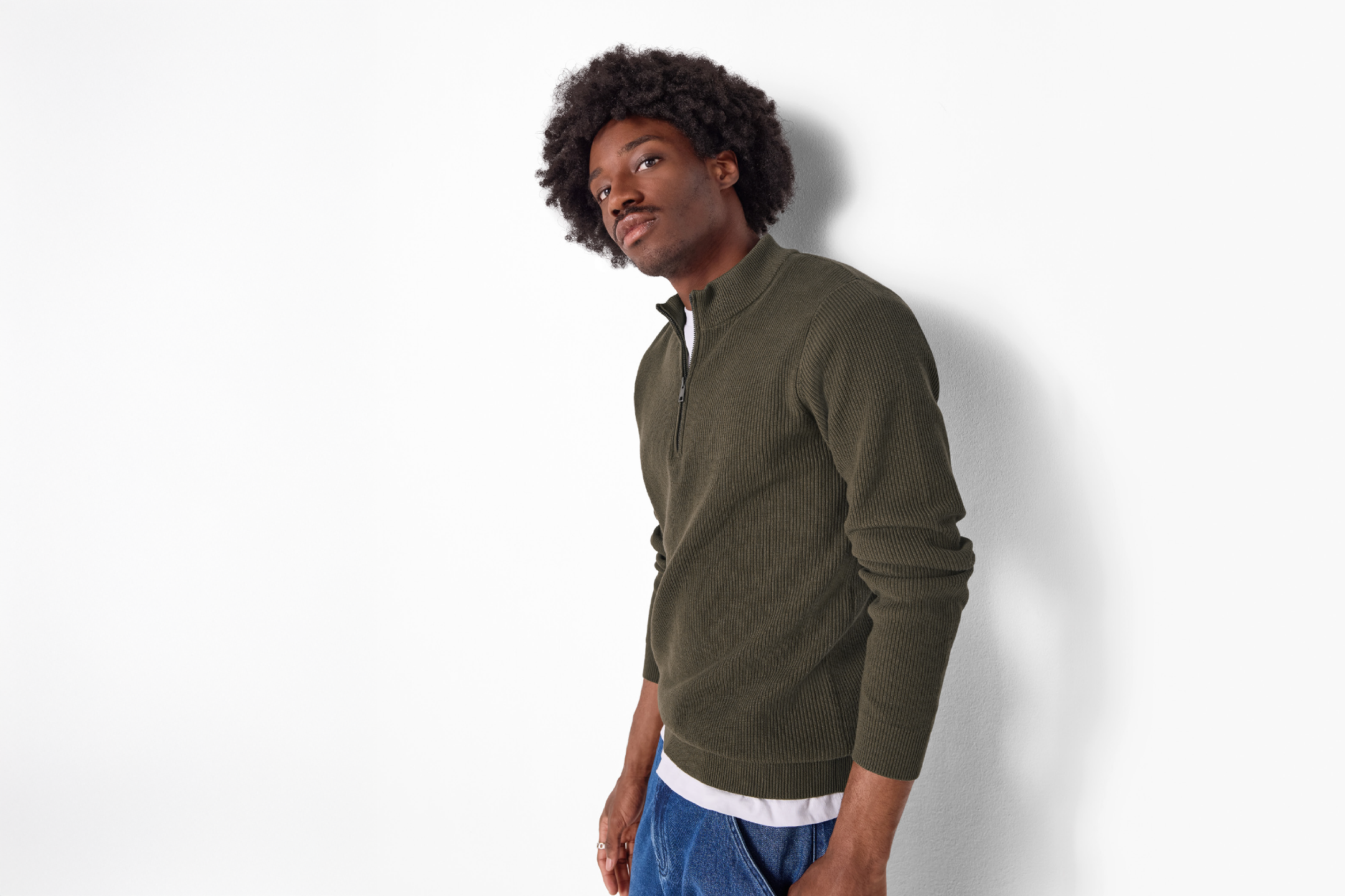 Man in a dark green ribbed quarter-zip sweater and blue jeans, leaning against a white wall.
