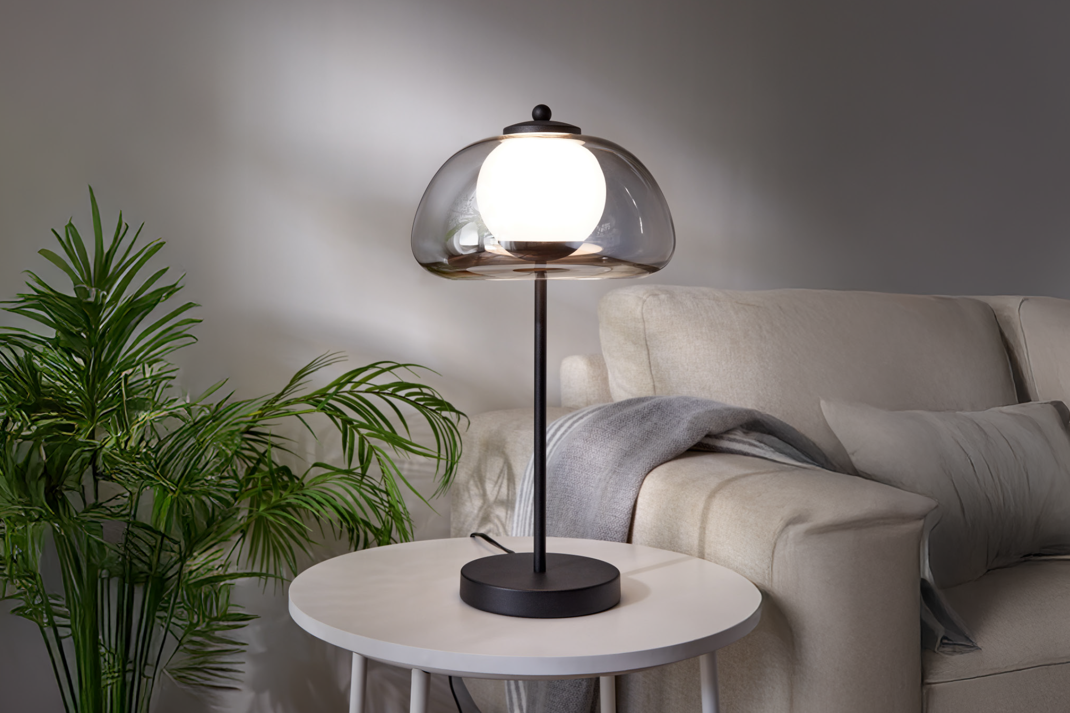 Modern table lamp with a smoked glass shade on a white side table next to a sofa and plant.