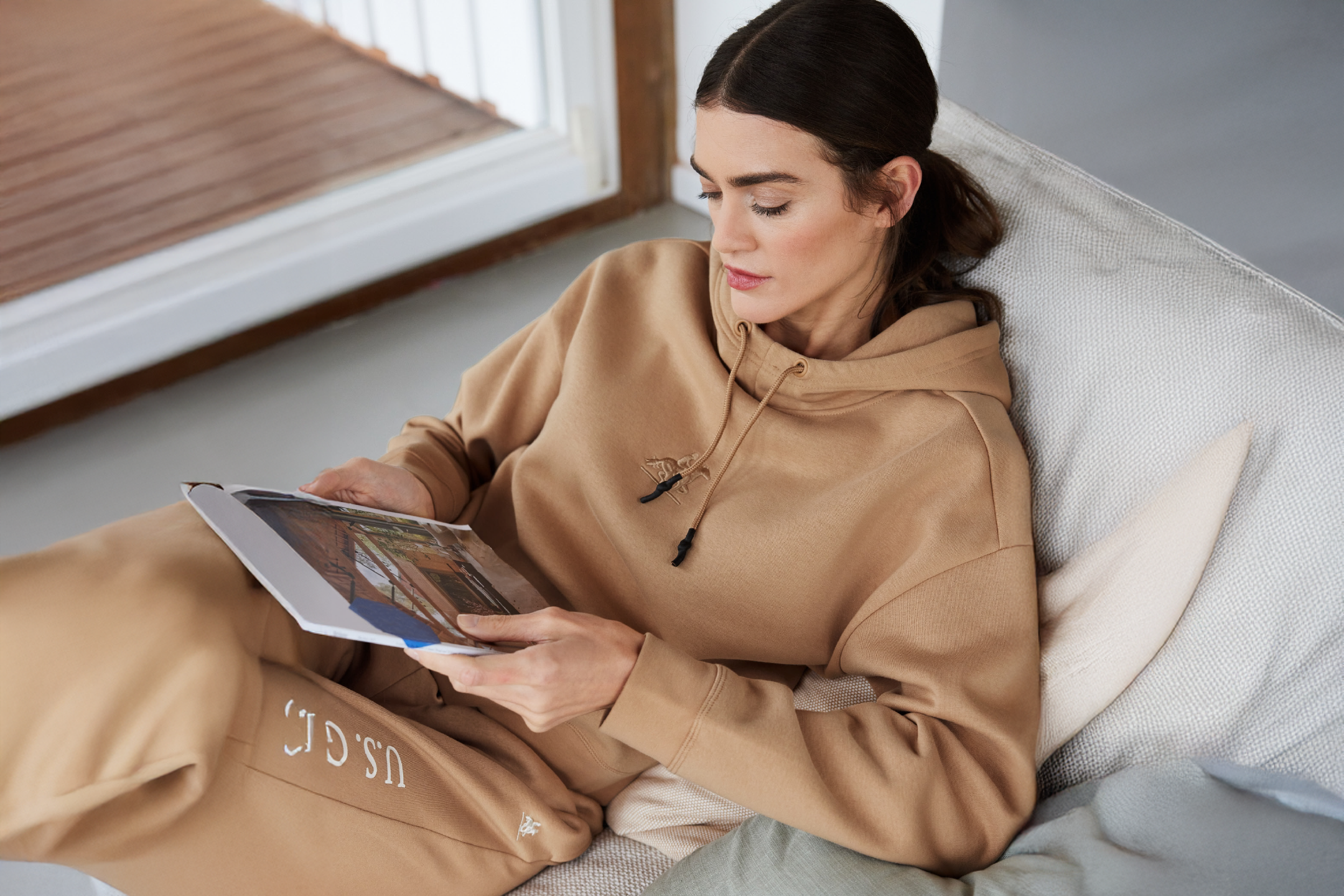 Woman in a beige hoodie and sweatpants reading a magazine on a sofa.