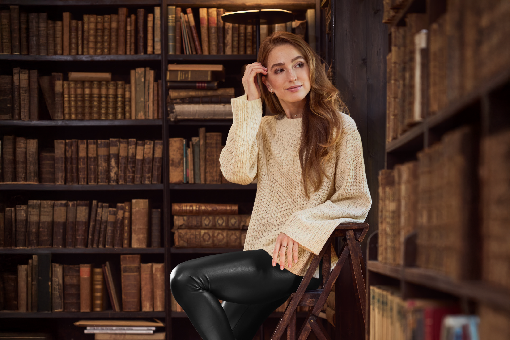 Woman in a cream sweater and black leggings sitting in a library.