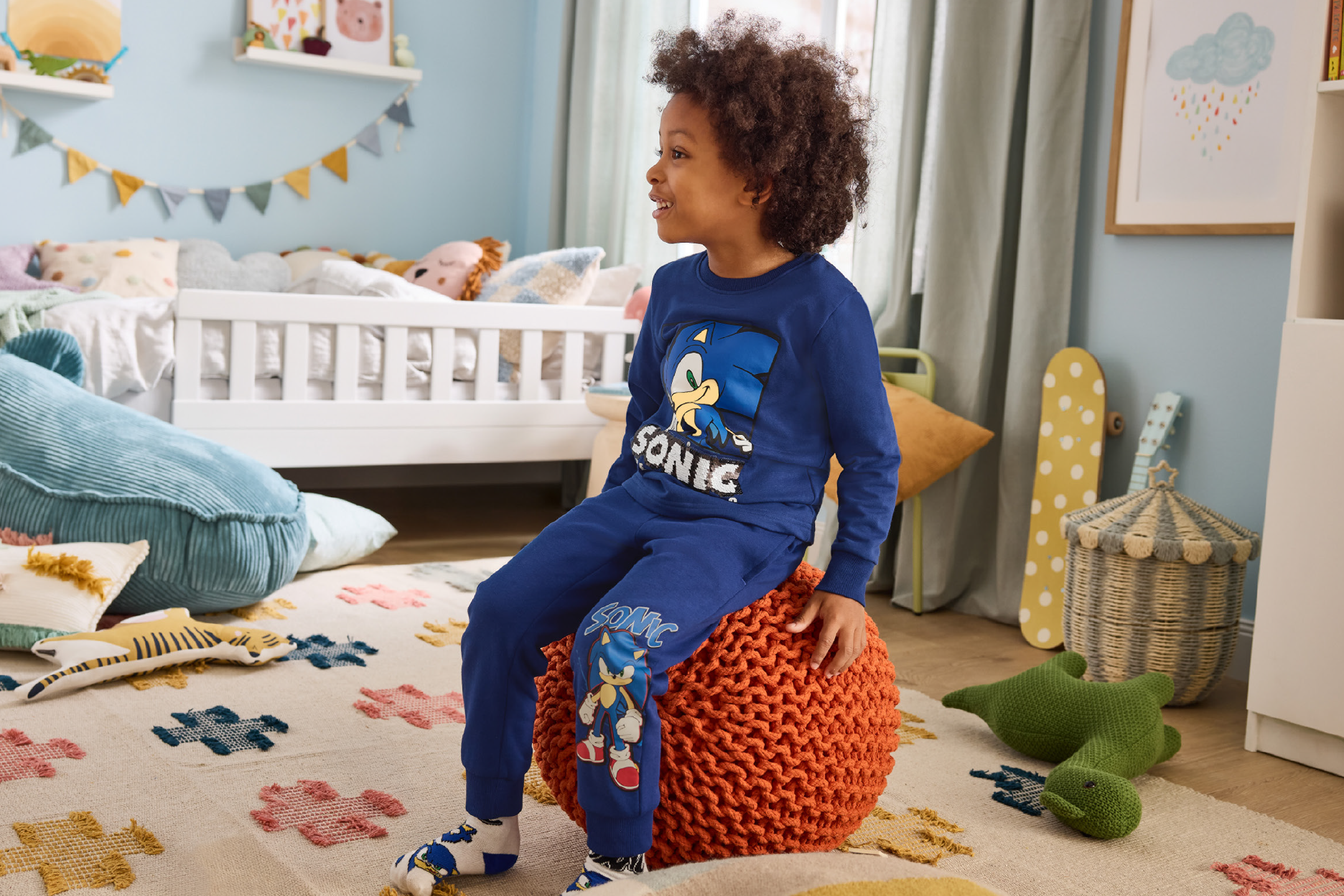 Child in blue Sonic the Hedgehog pajamas sitting on an orange pouf in a bedroom.
