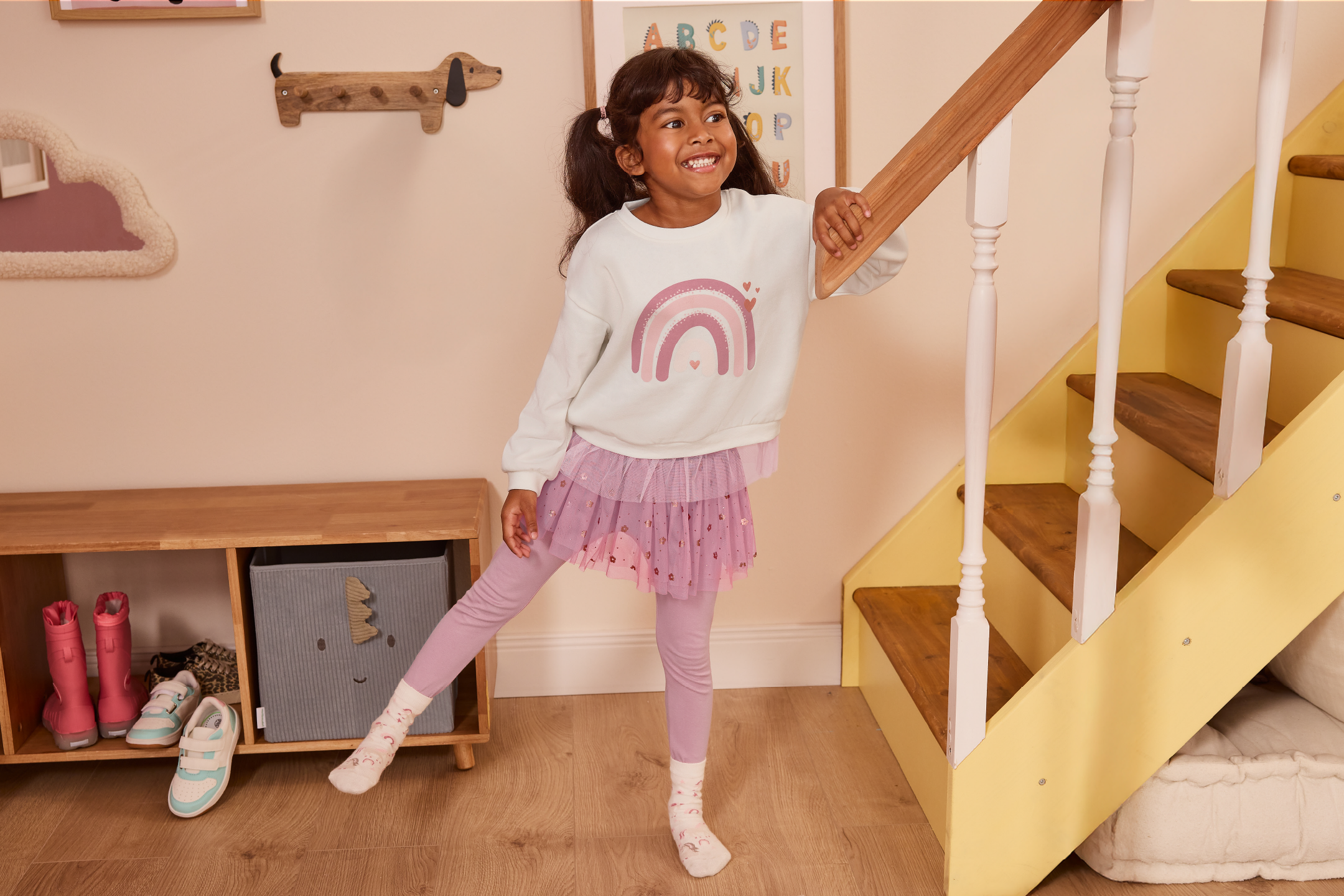 Girl in a rainbow sweatshirt and pink tulle skirt, next to a wooden staircase.