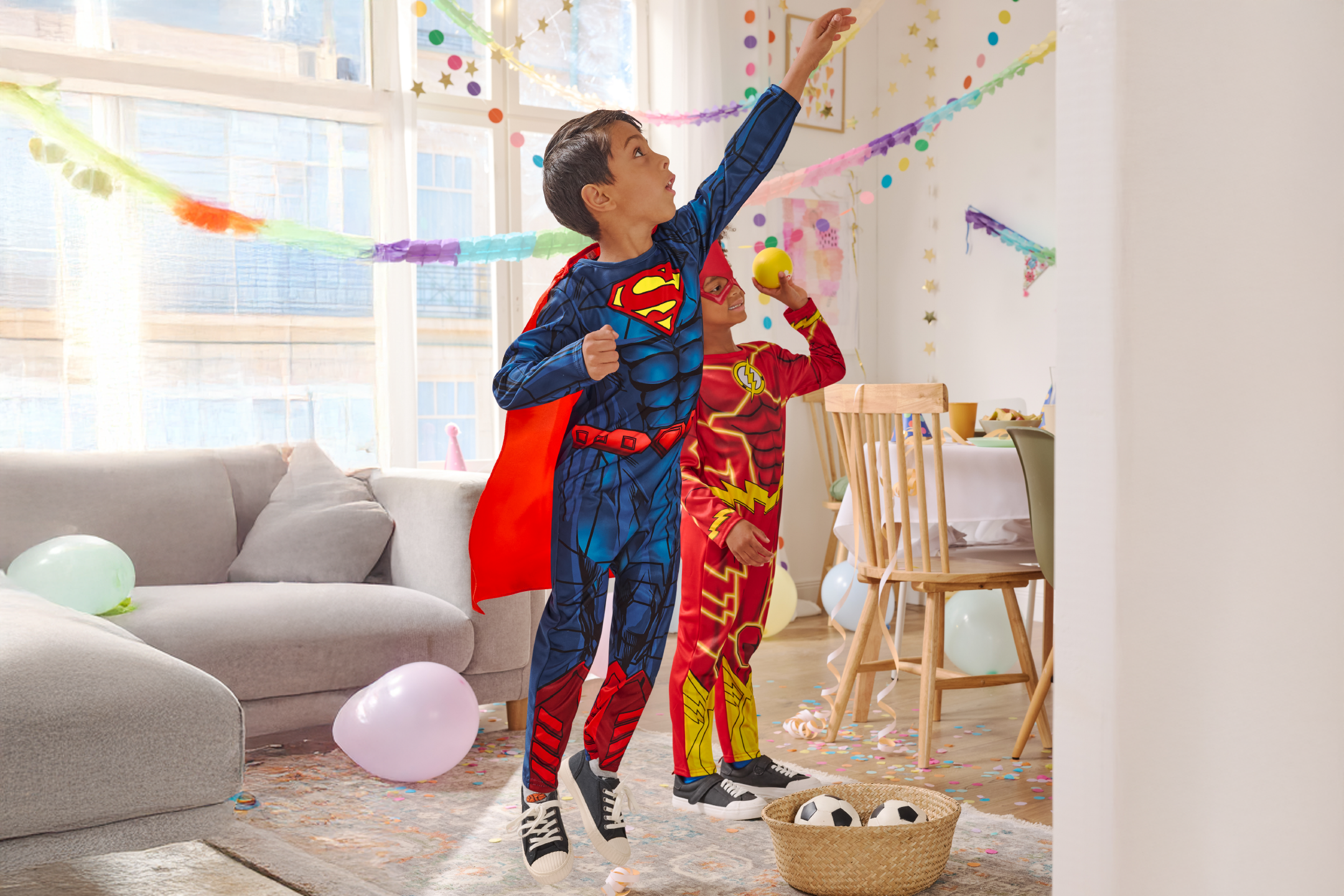 Two boys in superhero costumes playing at a party, surrounded by balloons and confetti.