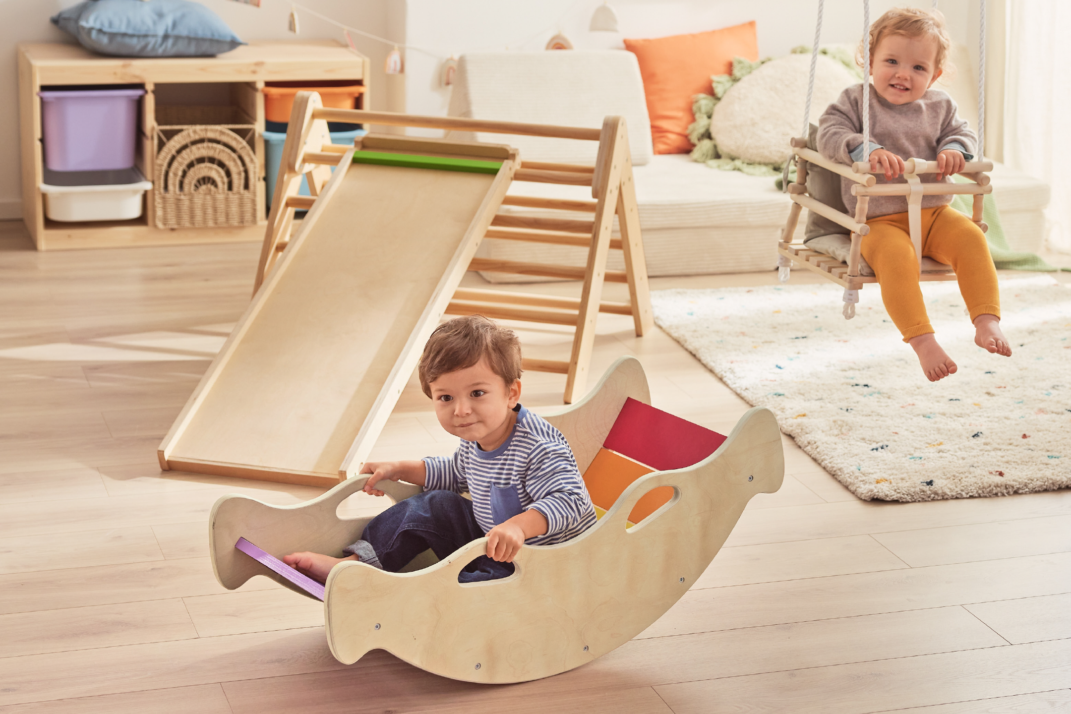 Two toddlers playing with a wooden slide, a wooden rocker, and a wooden swing in a bright room.