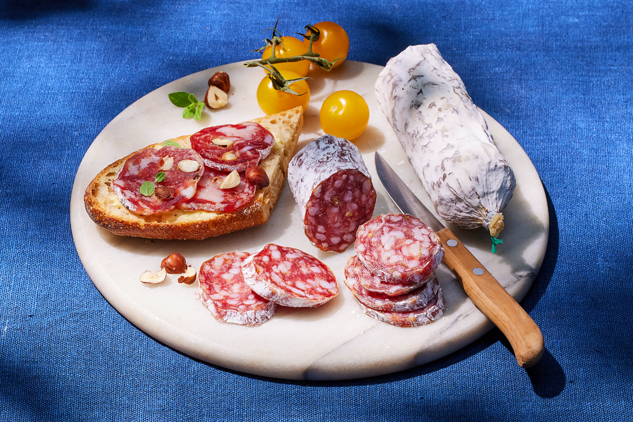 Φέτες και ολόκληρο σαλάμι σε μαρμάρινη σανίδα με ψωμί, ντομάτες και φουντούκια.