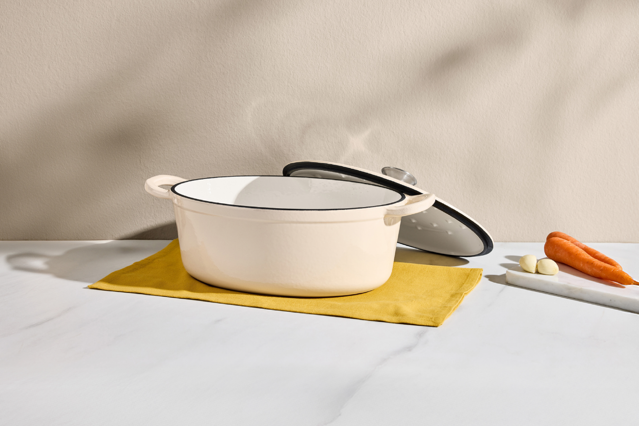 Κρεμ κατσαρόλα εμαγιέ με καπάκι, κίτρινο ύφασμα, καρότα και σκόρδο σε μαρμάρινη επιφάνεια.