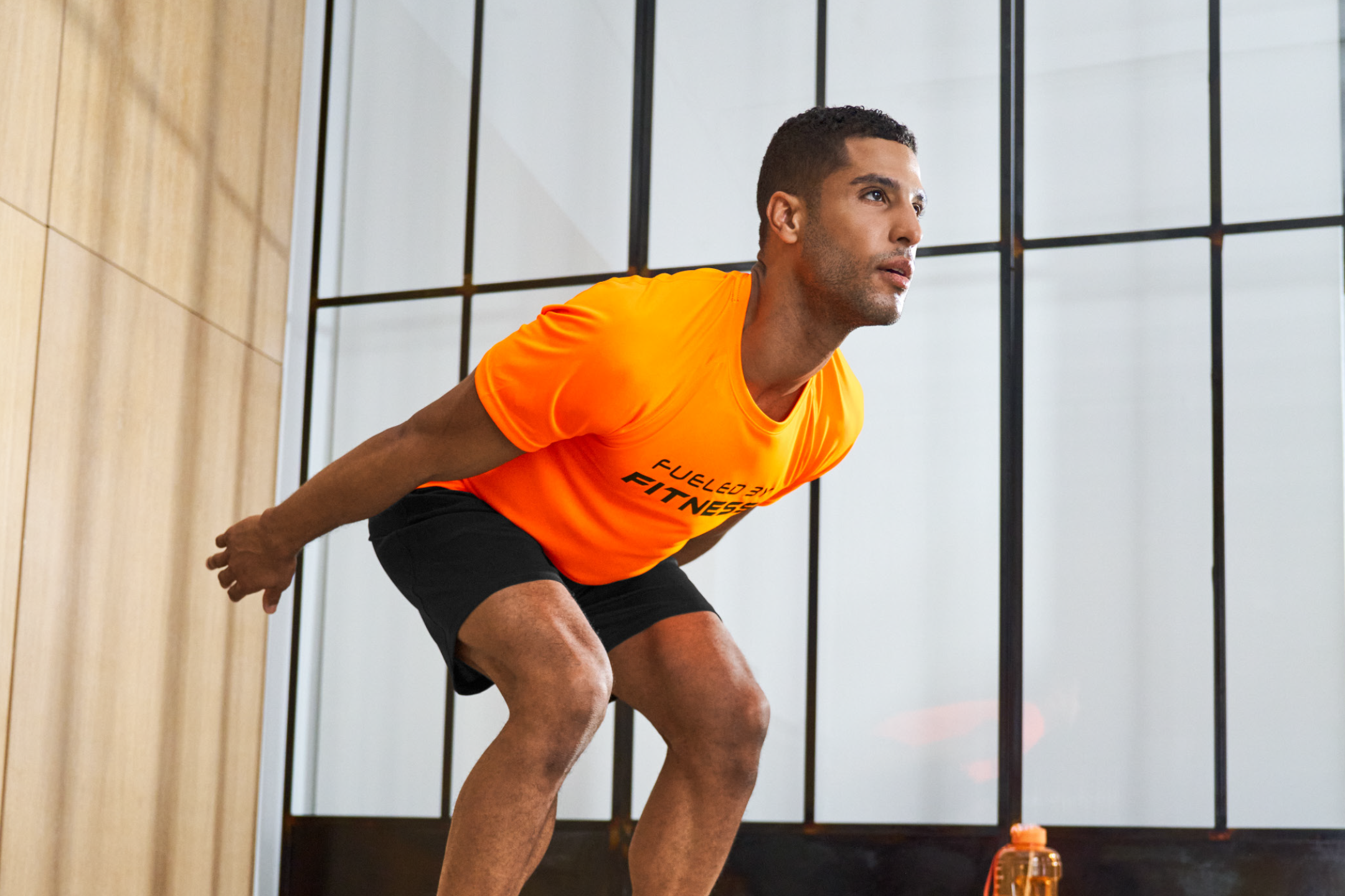 Man in orange fitness t-shirt and black shorts doing squats, text 'FUELED BY FITNESS'