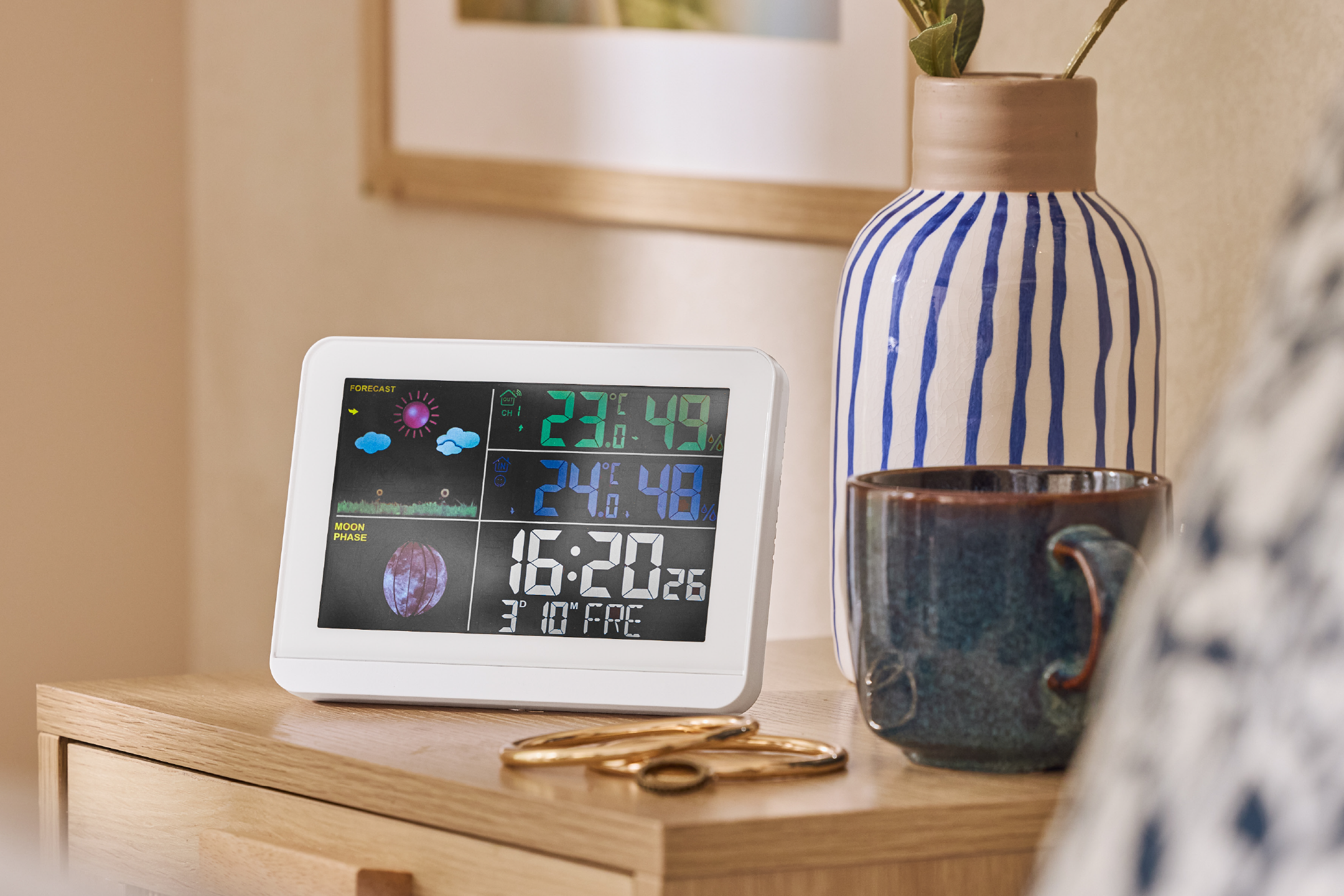 Digital weather station displaying forecast, temperature, humidity, moon phase, and time on a bedside table.
