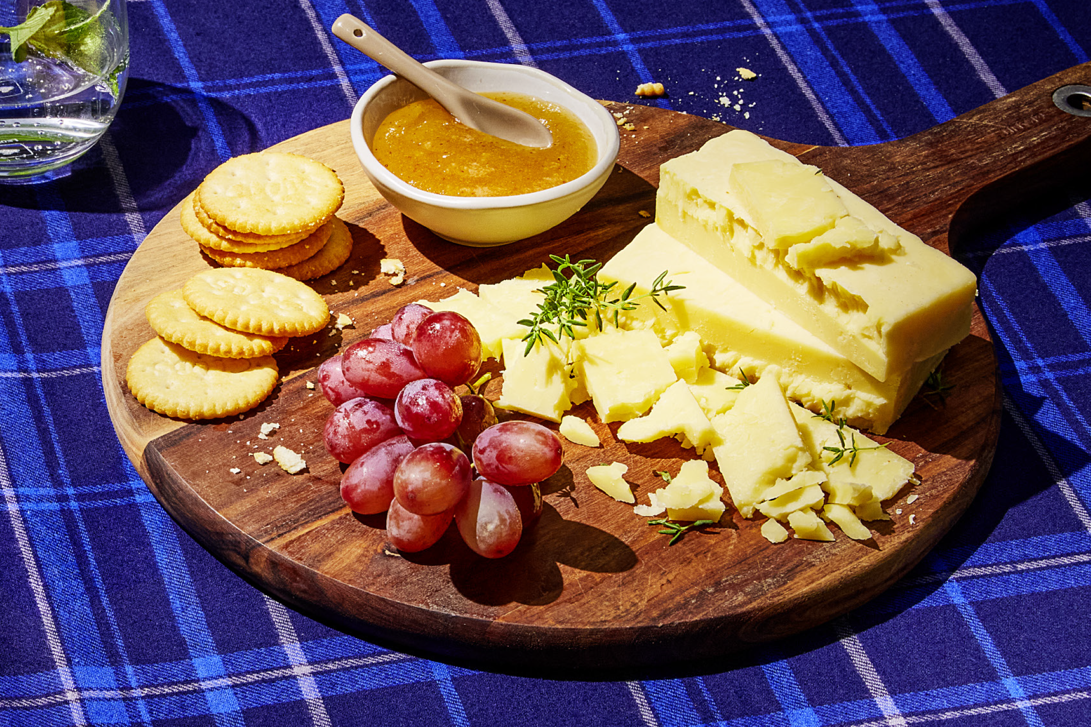 Cheese board with crackers, grapes, and jam on a blue plaid tablecloth.