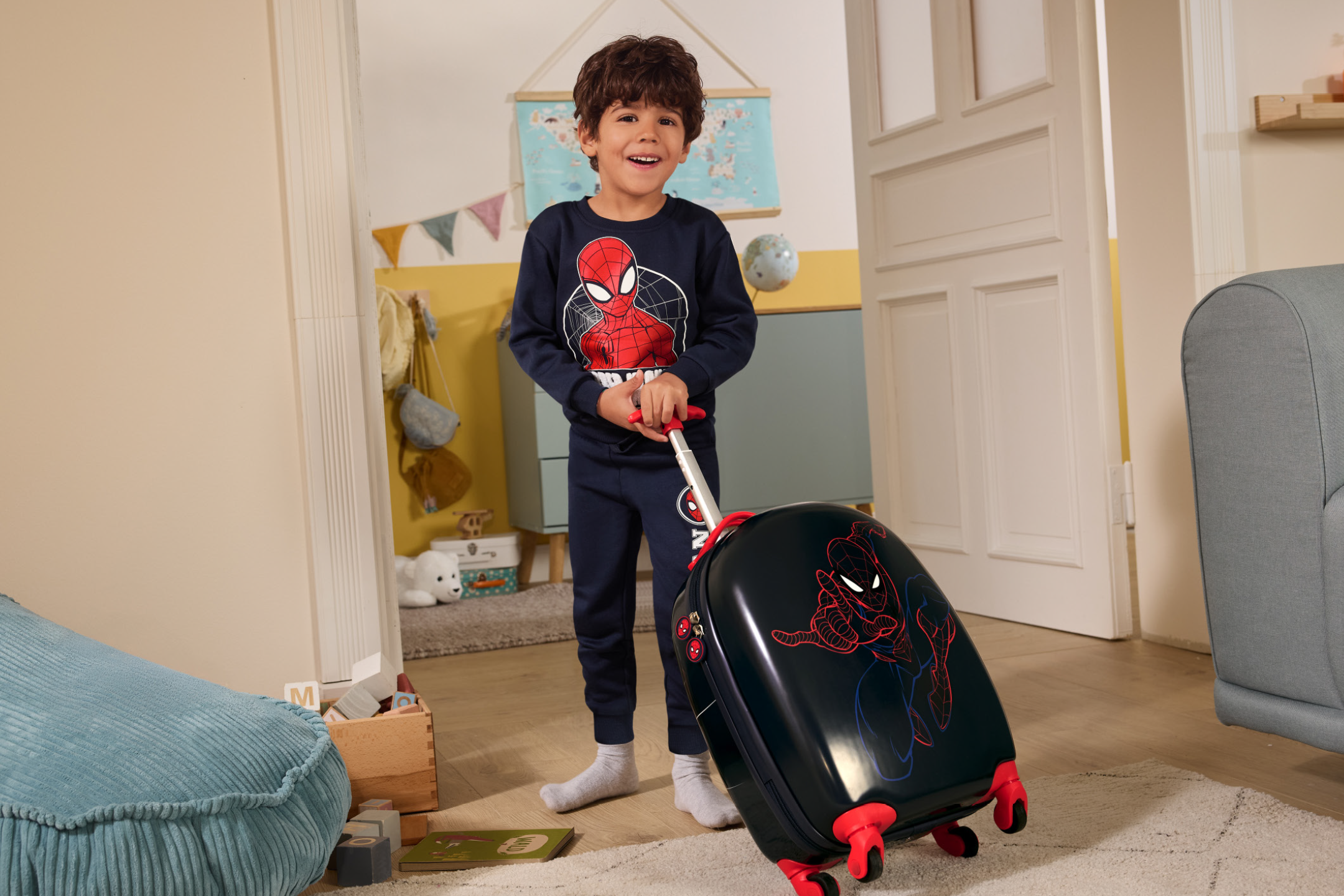 Smiling boy in a blue long-sleeve top and trousers, pulling a black suitcase with a superhero design.