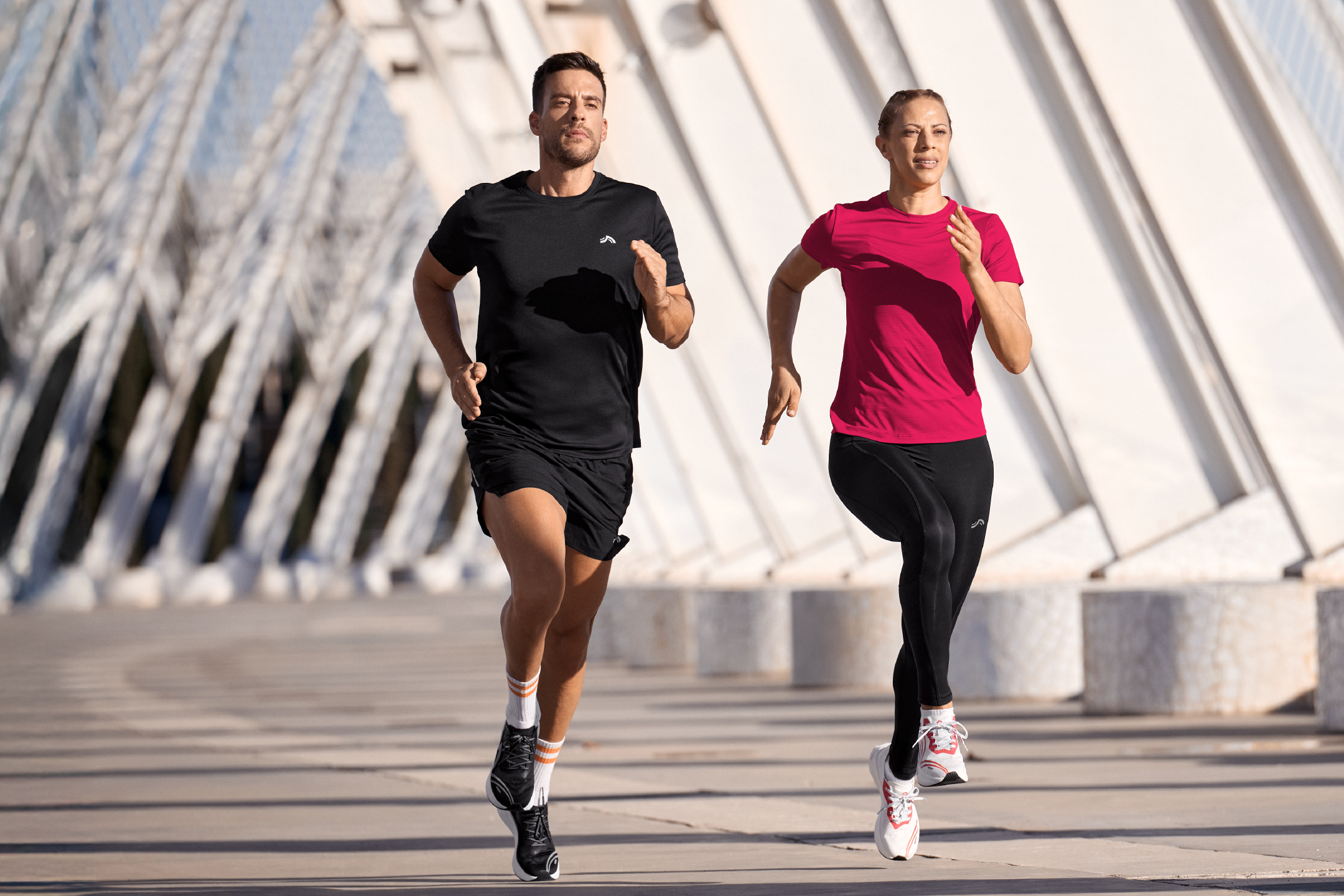 Two people running outdoors, a man in a black t-shirt and shorts, a woman in a pink t-shirt and black leggings.