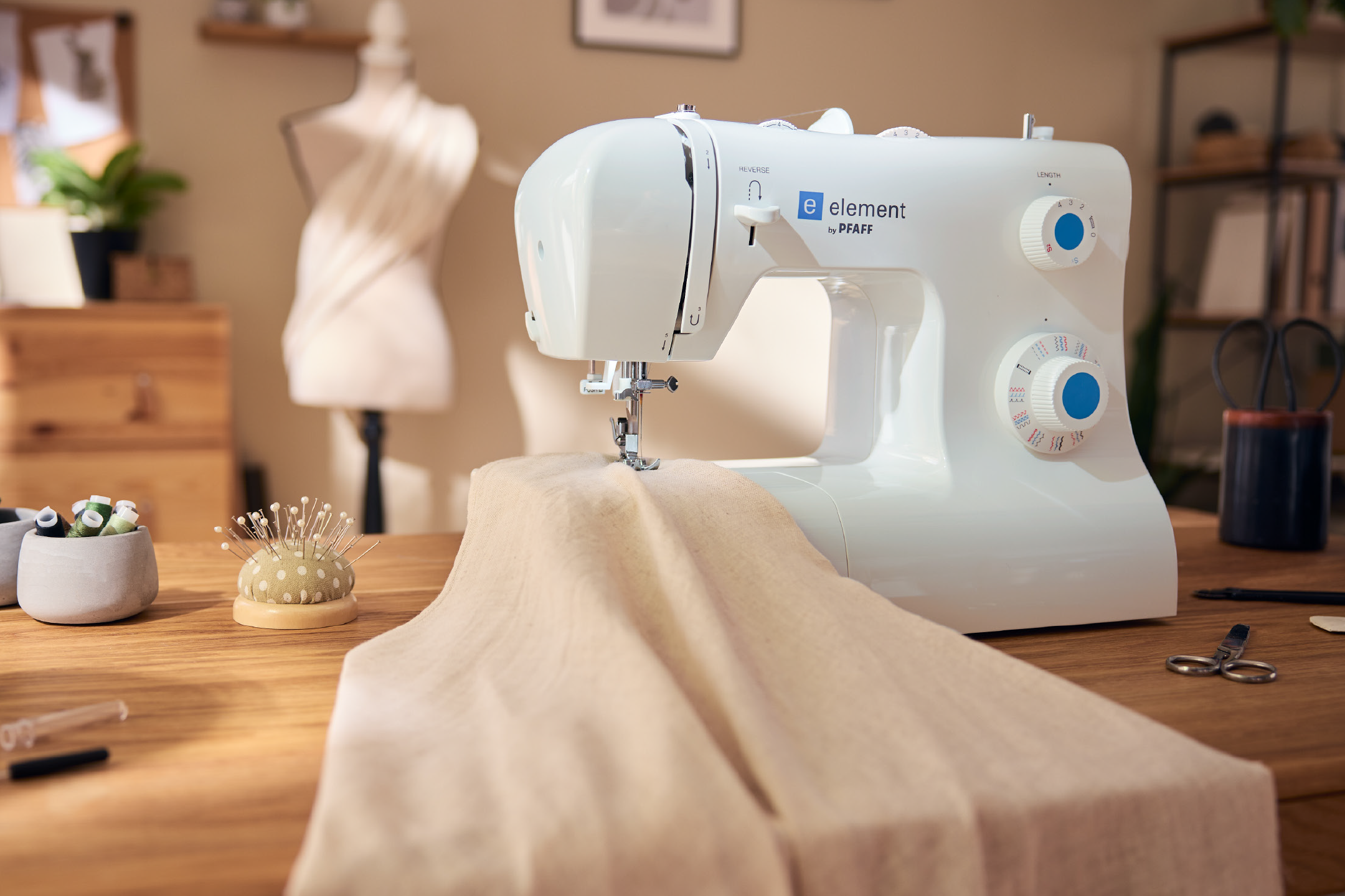 Λευκή ραπτομηχανή με ύφασμα, μαξιλαράκι για καρφίτσες και κλωστές σε ξύλινο τραπέζι.