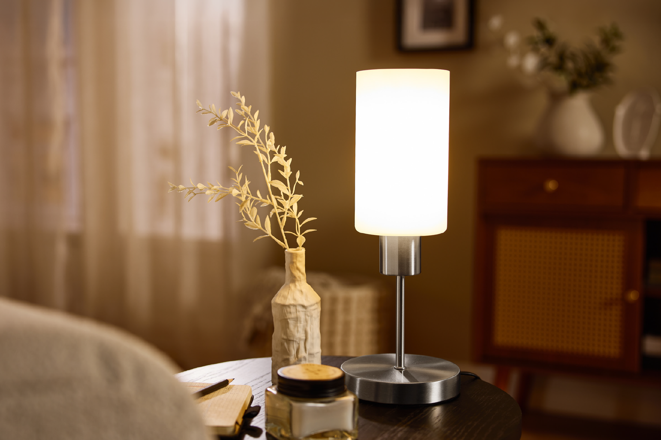 Επιτραπέζιο φωτιστικό με διακοσμητικό βάζο και αποξηραμένο φυτό σε βοηθητικό τραπέζι σε ένα ζεστό δωμάτιο.