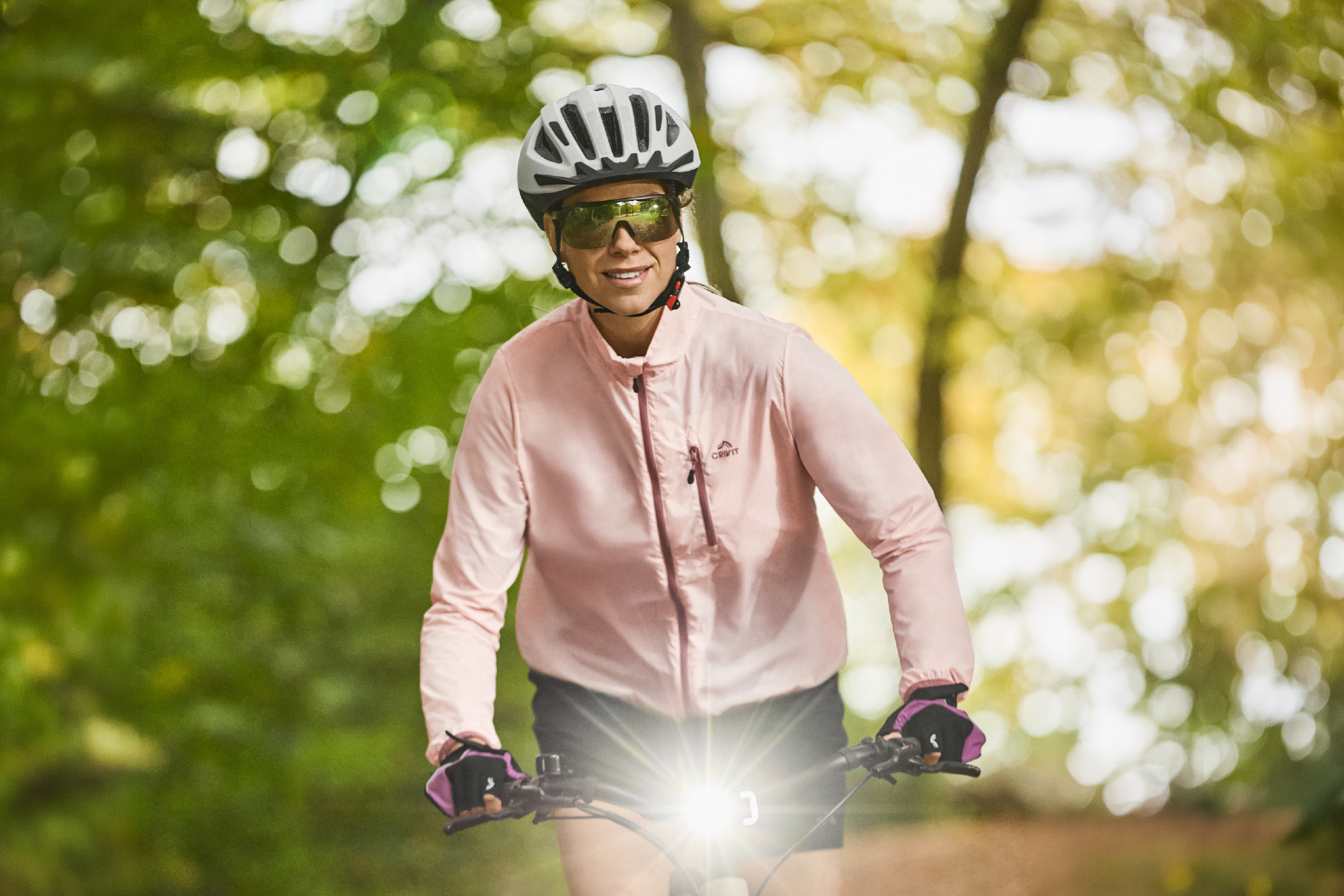 Γυναίκα με ροζ ποδηλατικό μπουφάν, κράνος και γυαλιά ηλίου οδηγεί ποδήλατο με έντονο φως.