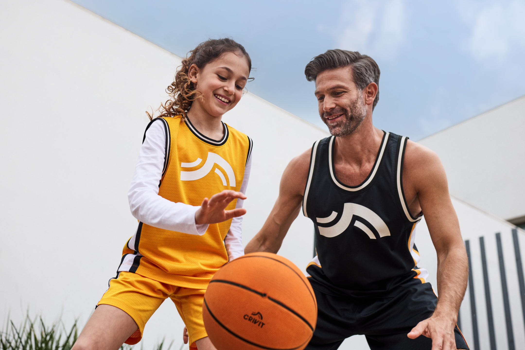 Πατέρας και κόρη παίζουν μπάσκετ, φορώντας κίτρινες και μαύρες φανέλες και σορτς μπάσκετ.