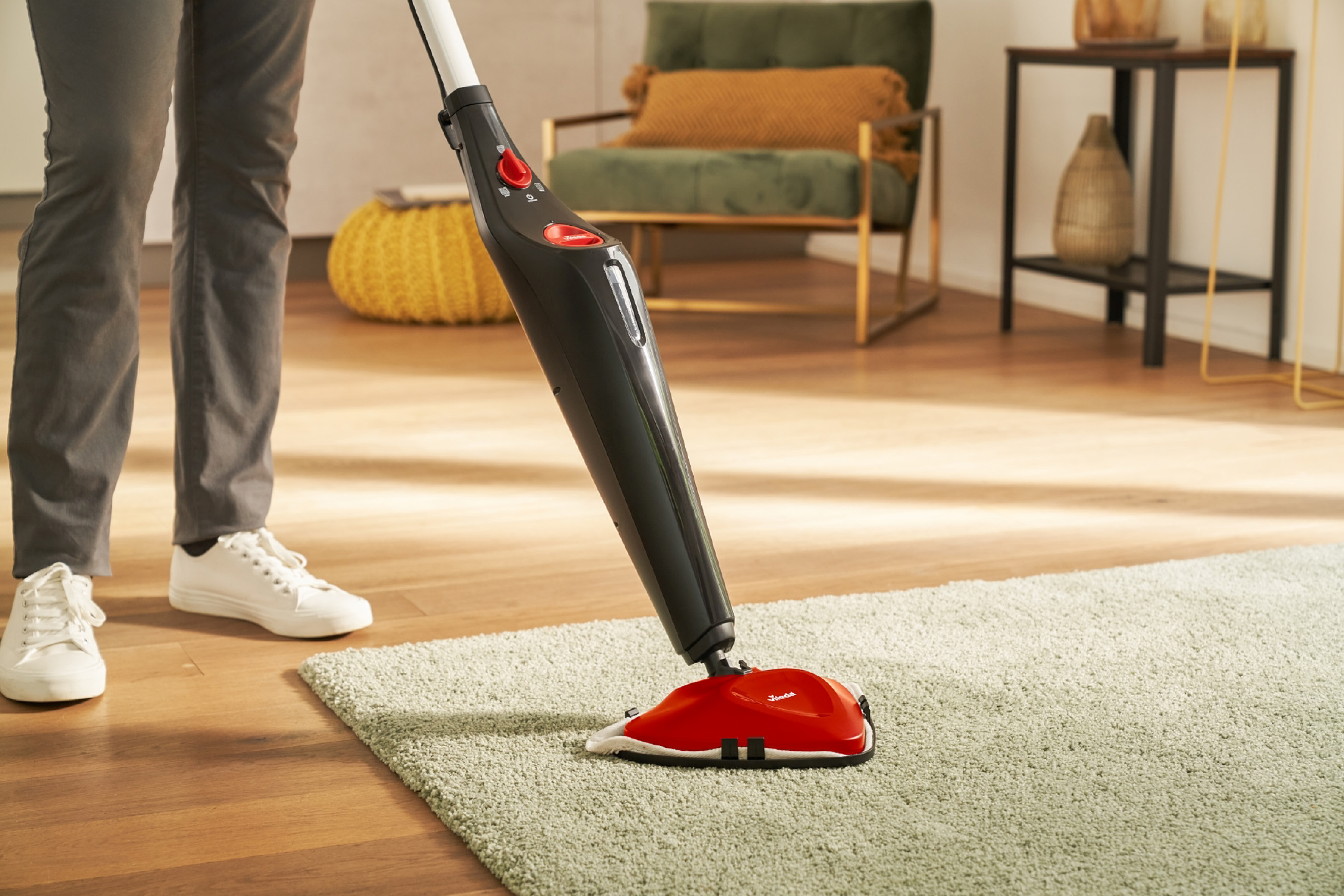 Person cleaning a light green rug with a steam mop in a living room.