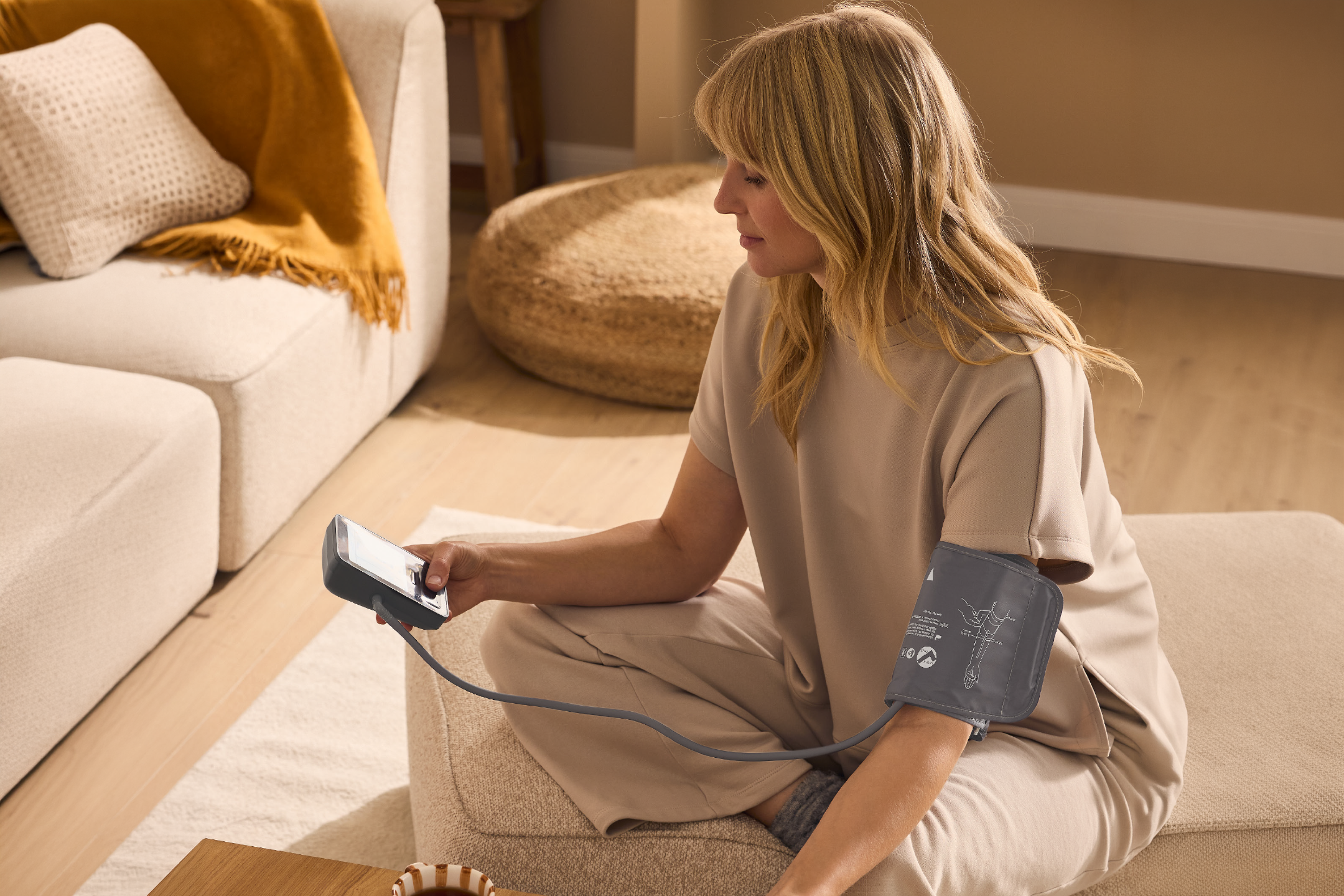 Woman measuring blood pressure at home, sitting on a beige floor cushion.