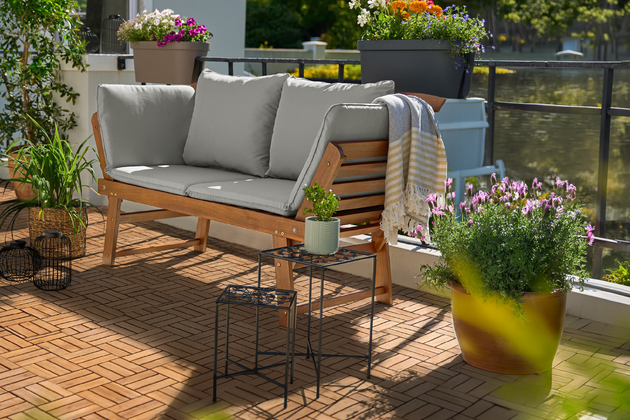 Outdoor wooden sofa with grey cushions, potted plants, and decorative items on a balcony.