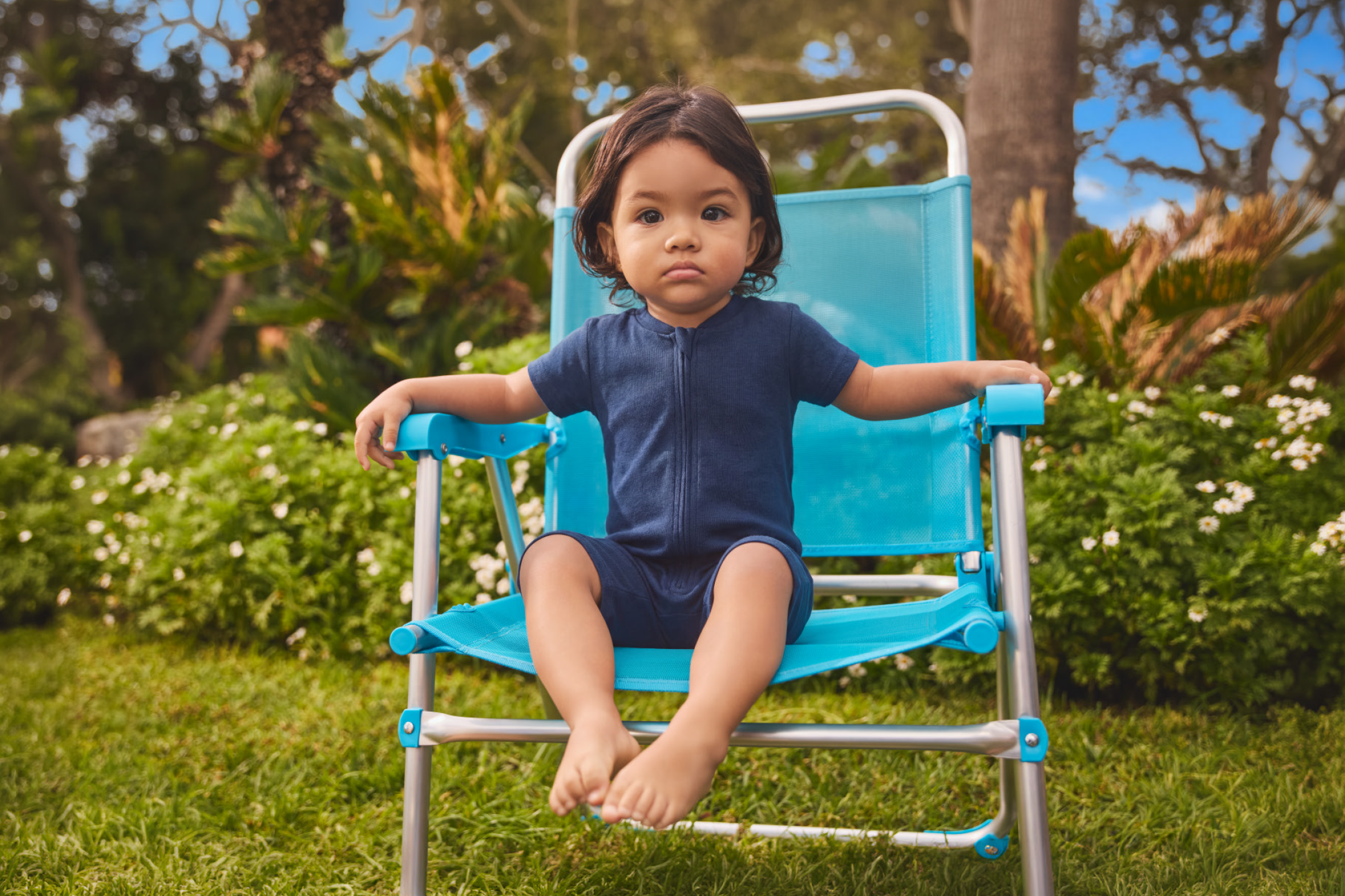 Toddler in a blue romper sitting on a blue beach chair in a garden.