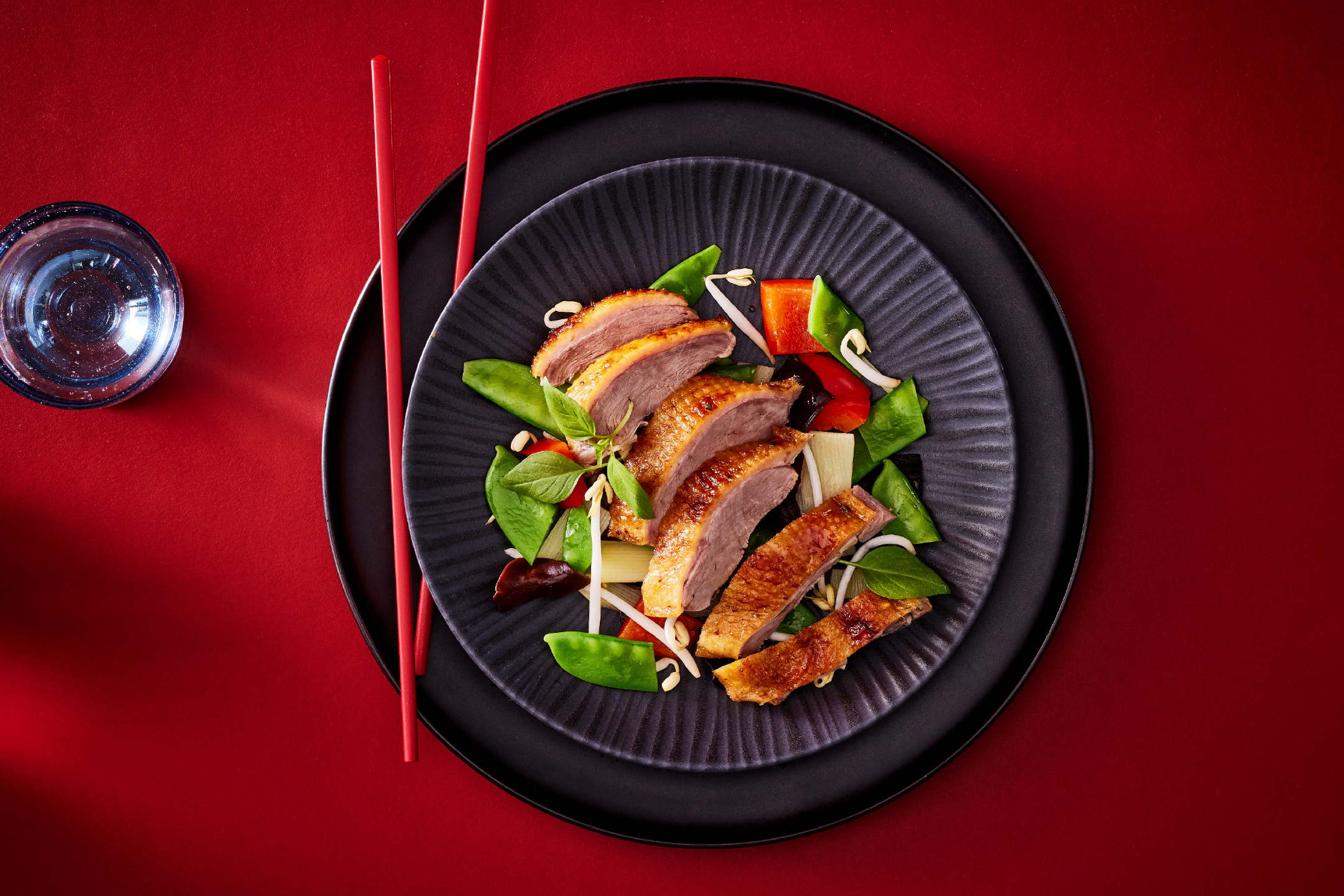 Crispy duck breast with stir-fried vegetables and bean sprouts on a black plate with red chopsticks.
