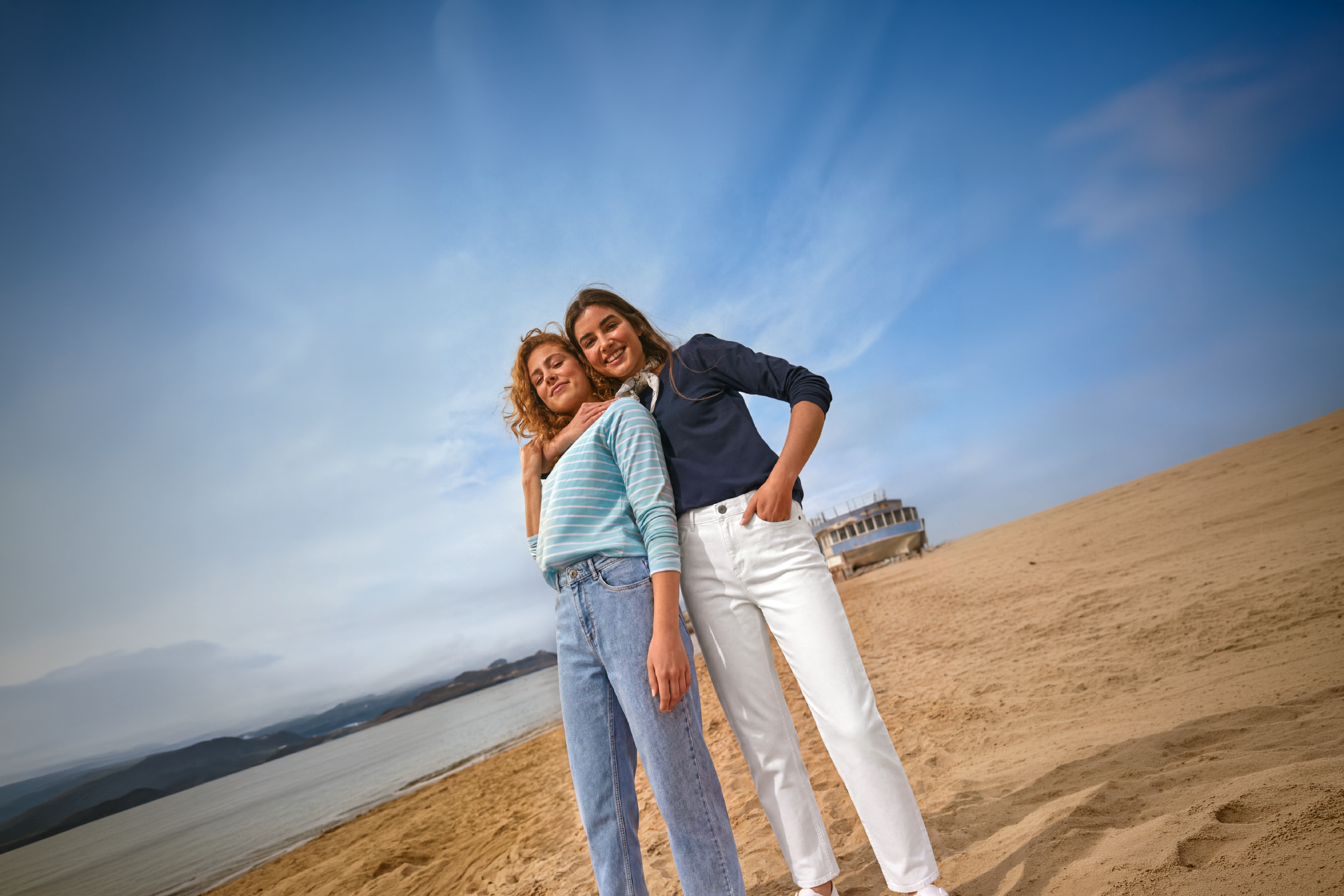 Two women in casual tops and jeans on a sandy beach with a boat in the background.