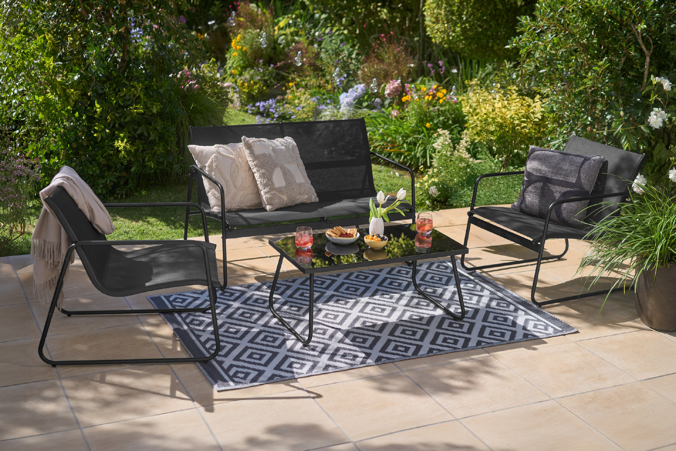 Σετ επίπλων εξωτερικού χώρου με μαύρο μεταλλικό σκελετό, που περιλαμβάνει διθέσιο καναπέ, δύο πολυθρόνες και τραπεζάκι σαλονιού, πάνω σε χαλί με σχέδια.