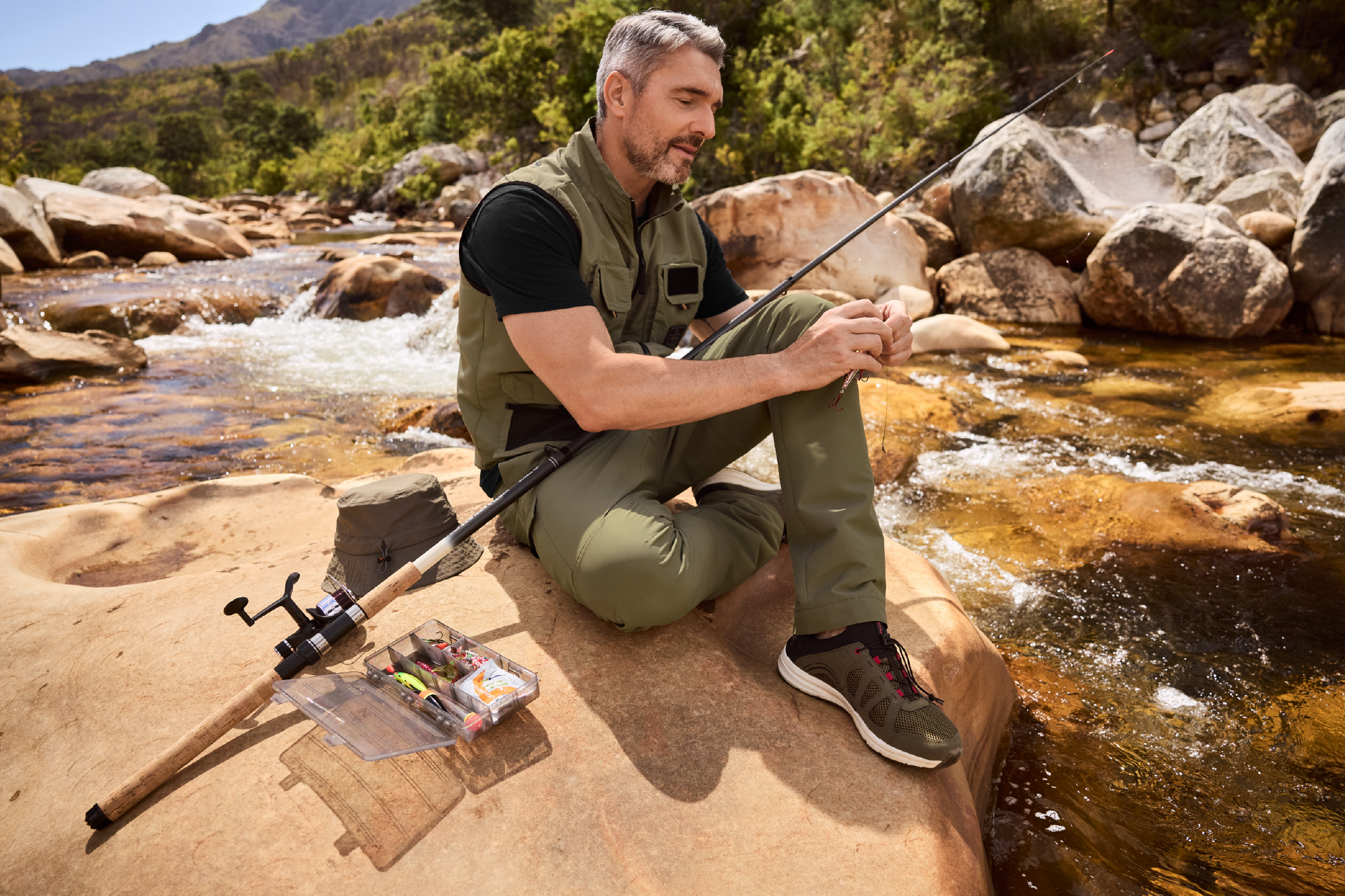 Άνδρας με γιλέκο και παντελόνι ψαρέματος, κάθεται δίπλα σε ποτάμι, ετοιμάζοντας τον εξοπλισμό ψαρέματος.