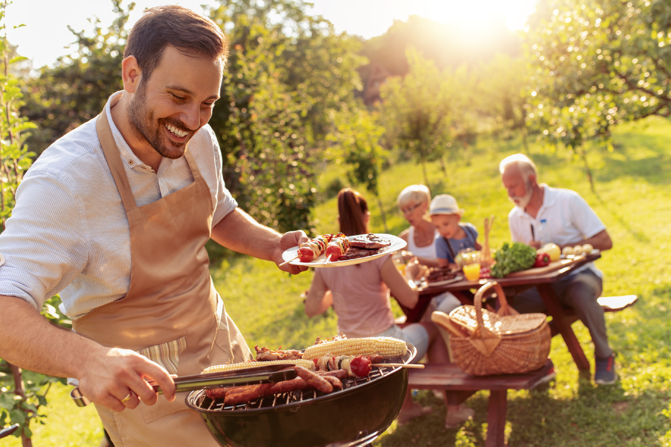 Άνδρας ψήνει λουκάνικα, καλαμπόκι και σουβλάκια σε οικογενειακό μπάρμπεκιου σε ηλιόλουστο κήπο.