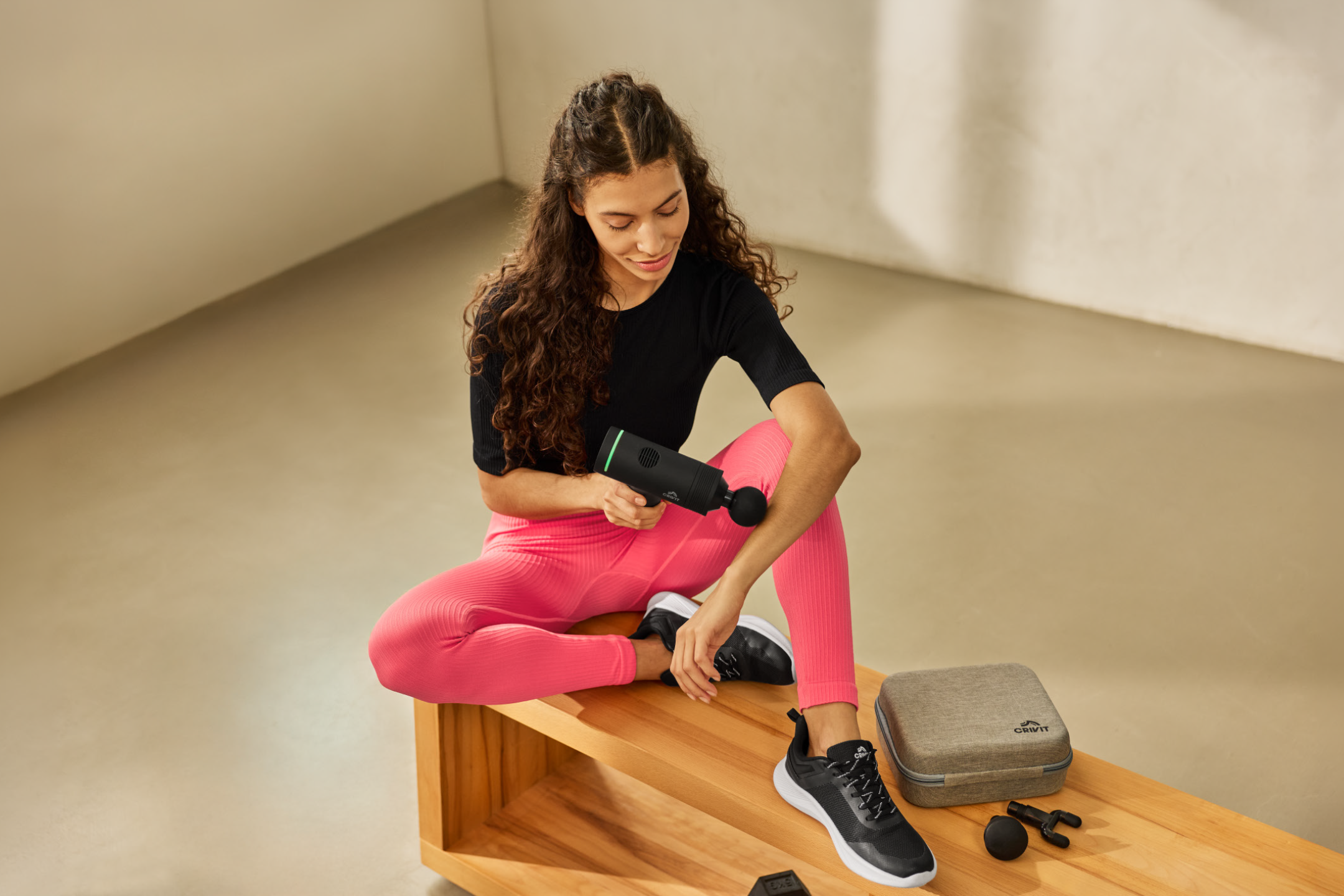 Γυναίκα με αθλητικά ρούχα χρησιμοποιεί πιστόλι μασάζ στο χέρι της, με μια θήκη δίπλα.