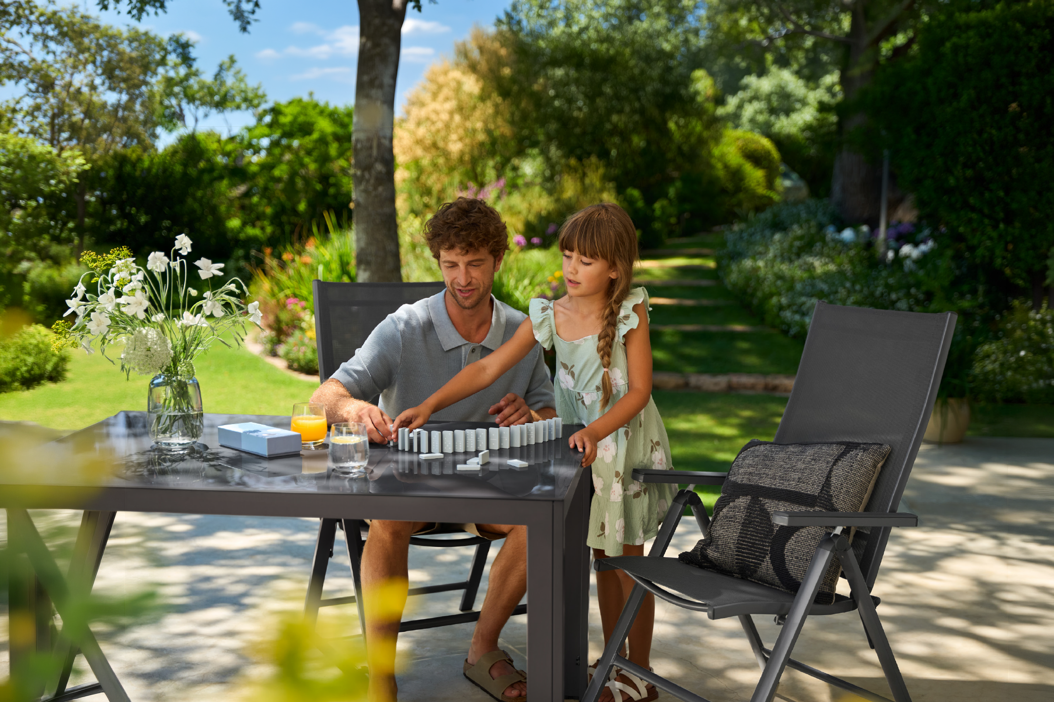 Father and daughter playing dominoes on a garden table with chairs in a sunny garden.