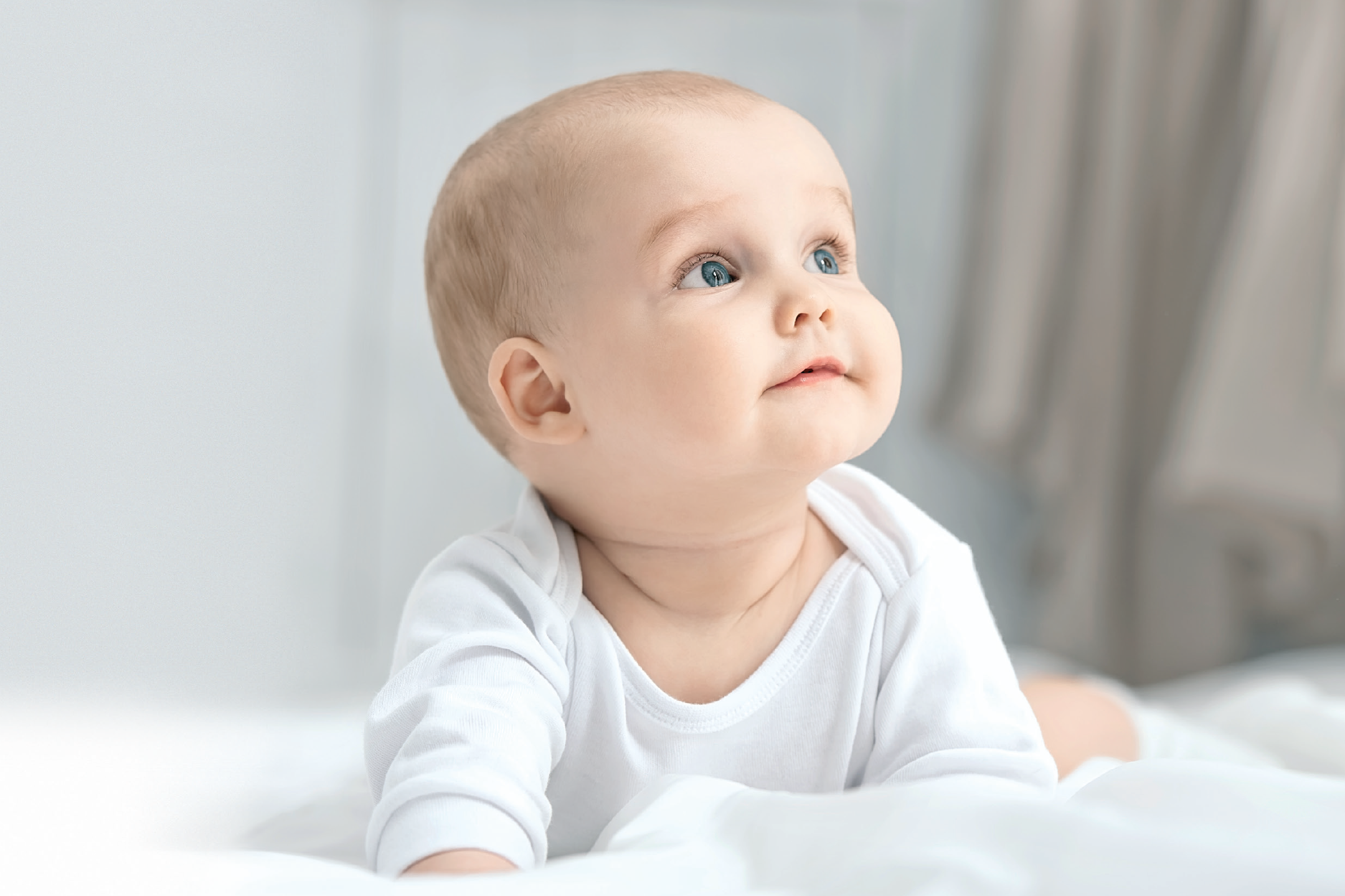 A baby in a white bodysuit lying on a white blanket, looking up with blue eyes.