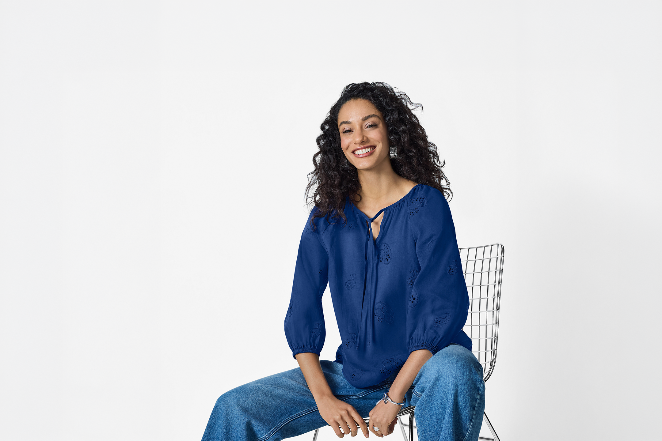Woman in a blue embroidered blouse and jeans, sitting on a metal chair and smiling.