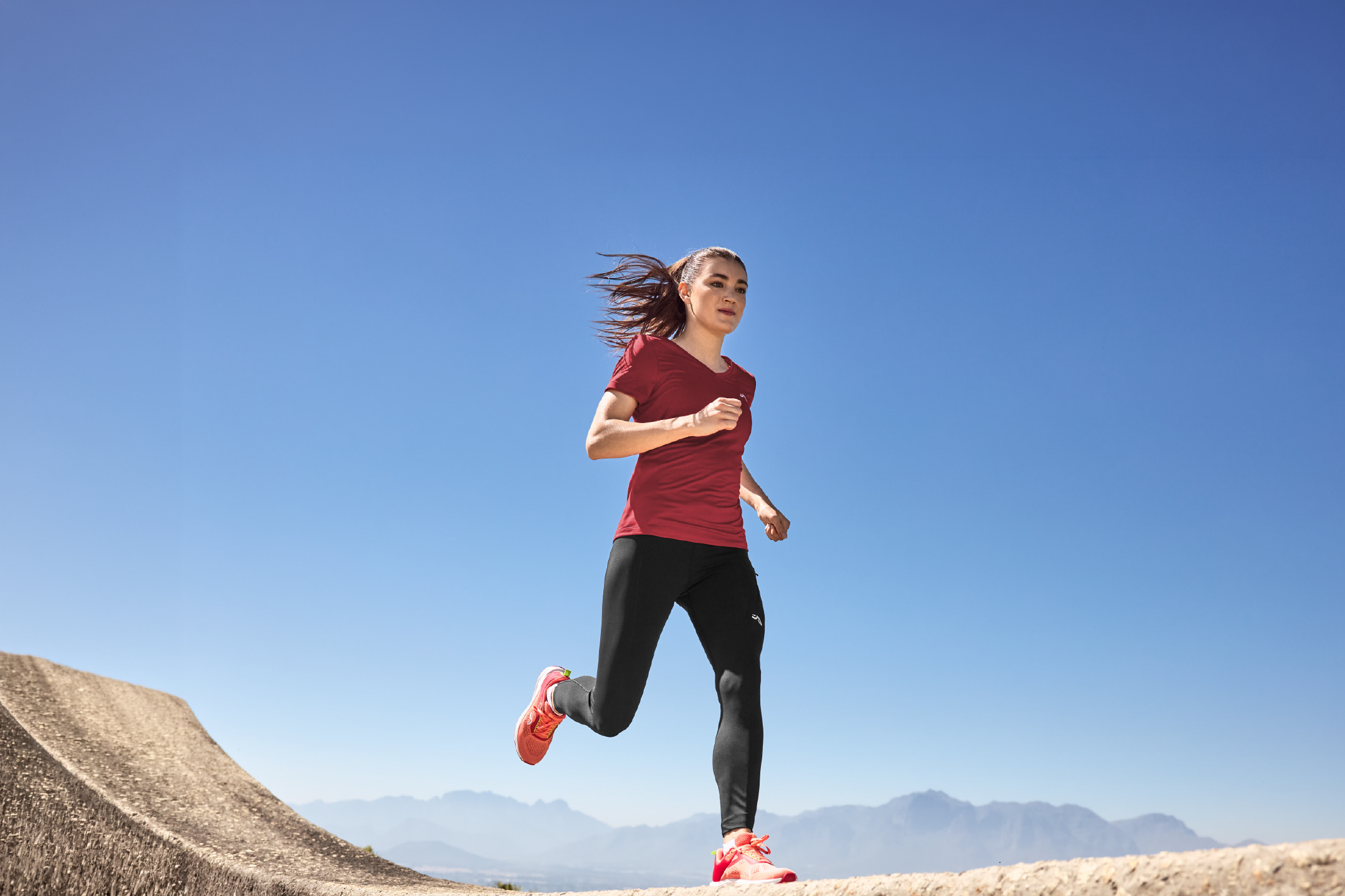 Woman in a red t-shirt and black leggings running outdoors with mountains in the background.
