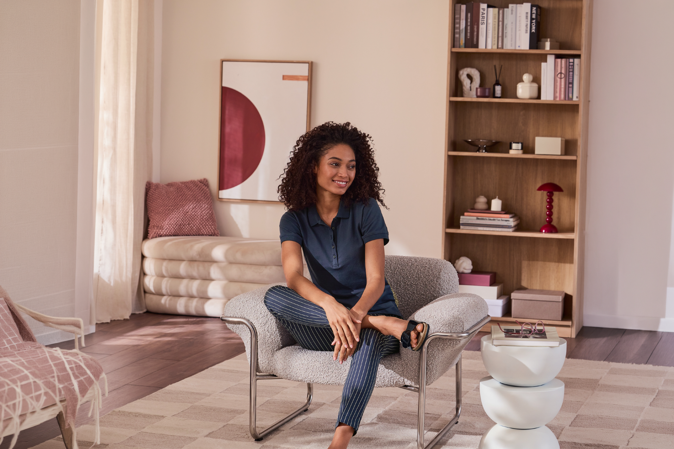 A woman in a blue polo shirt and striped pants sits in a living room with a bookshelf.