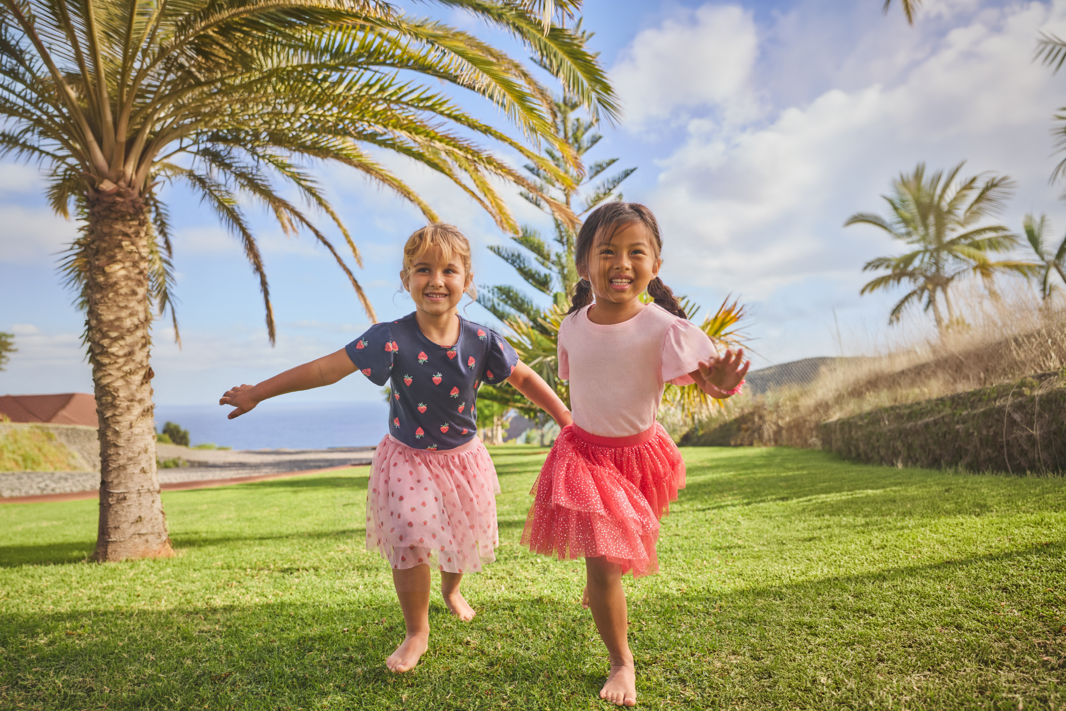 Two young girls in patterned skirts and t-shirts running barefoot on grass under palm trees.